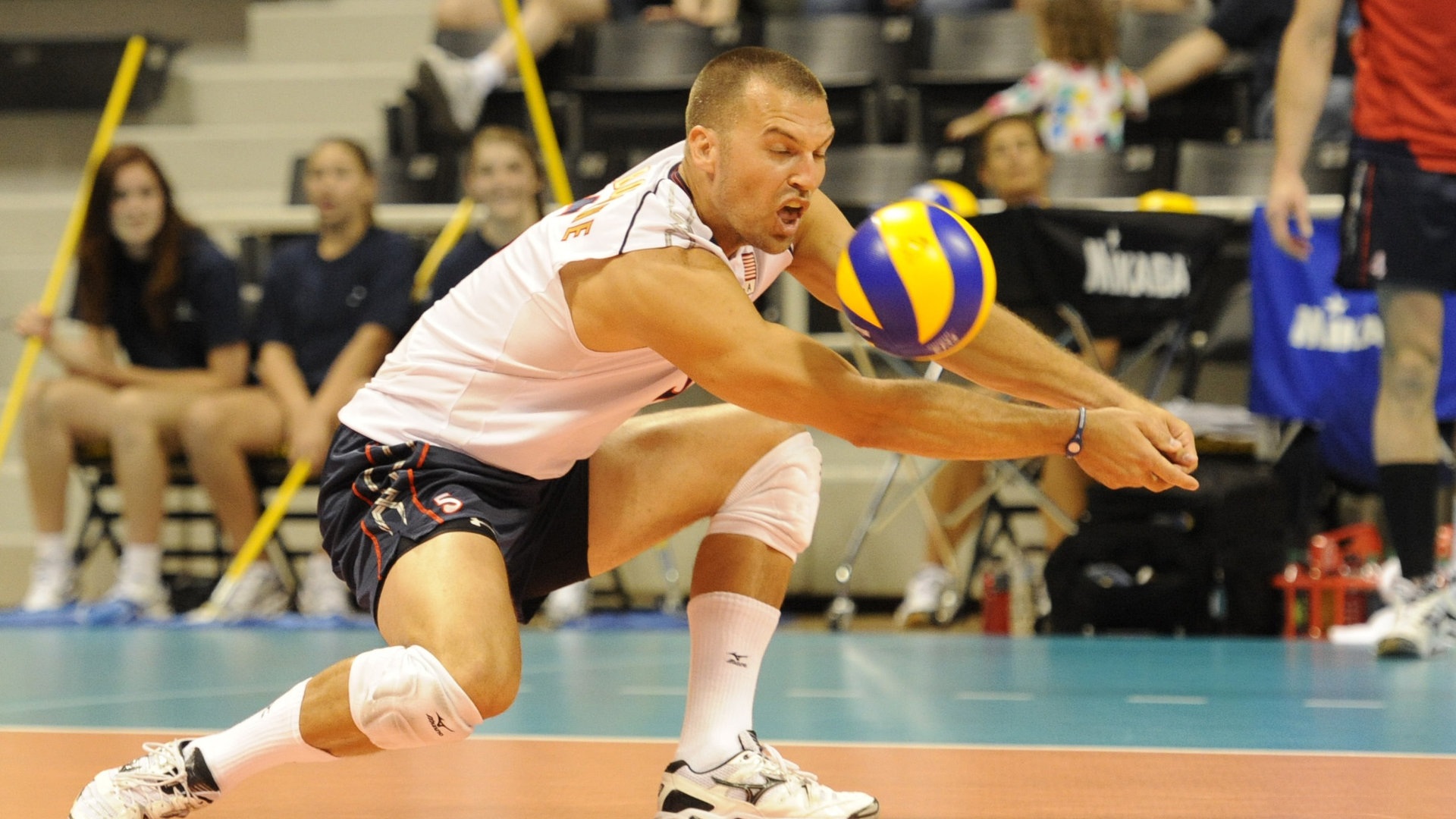 A male volleyball player in a white and blue uniform dives to receive the ball during a game