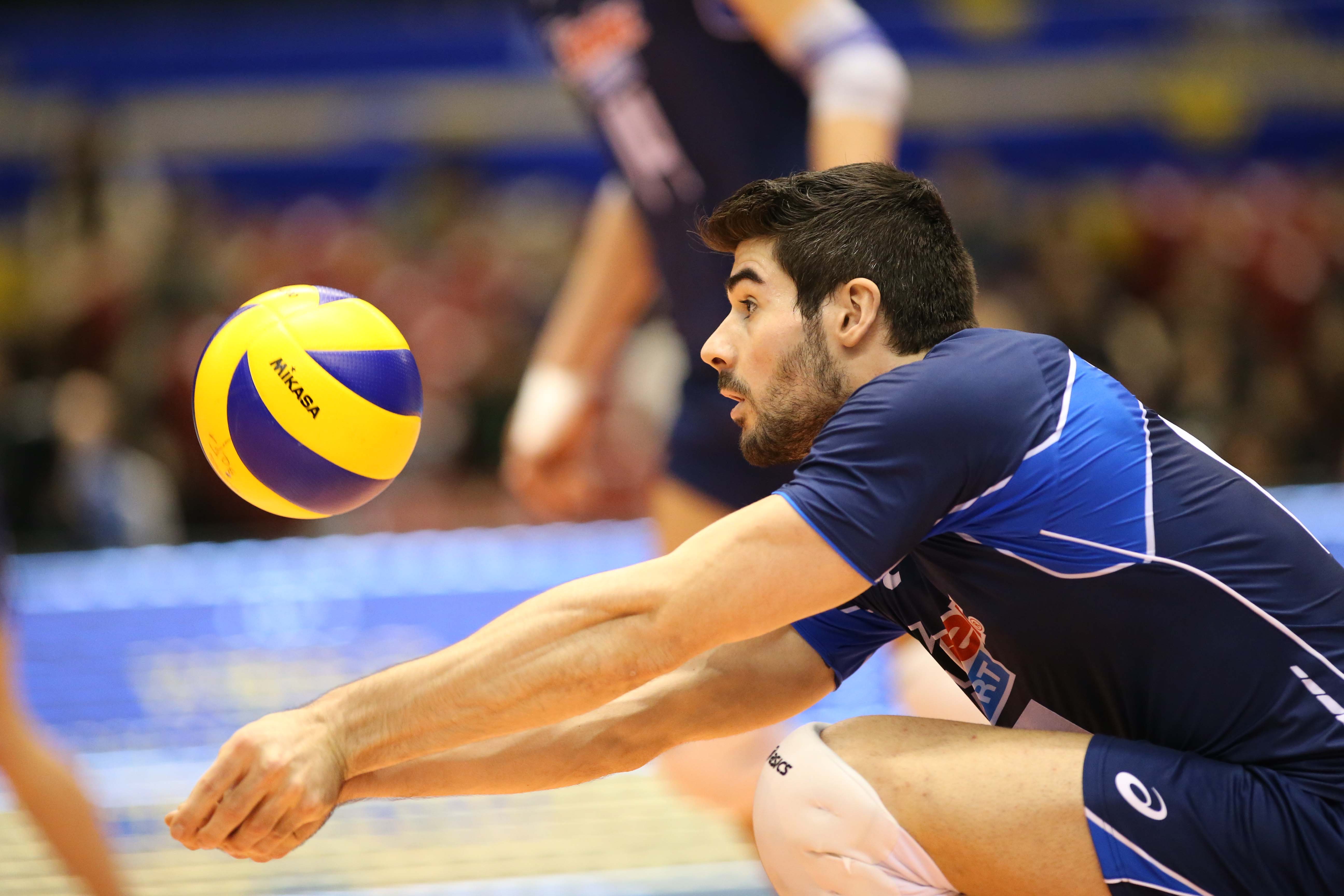 A male volleyball player in a blue and white uniform prepares to receive the ball during a professional match