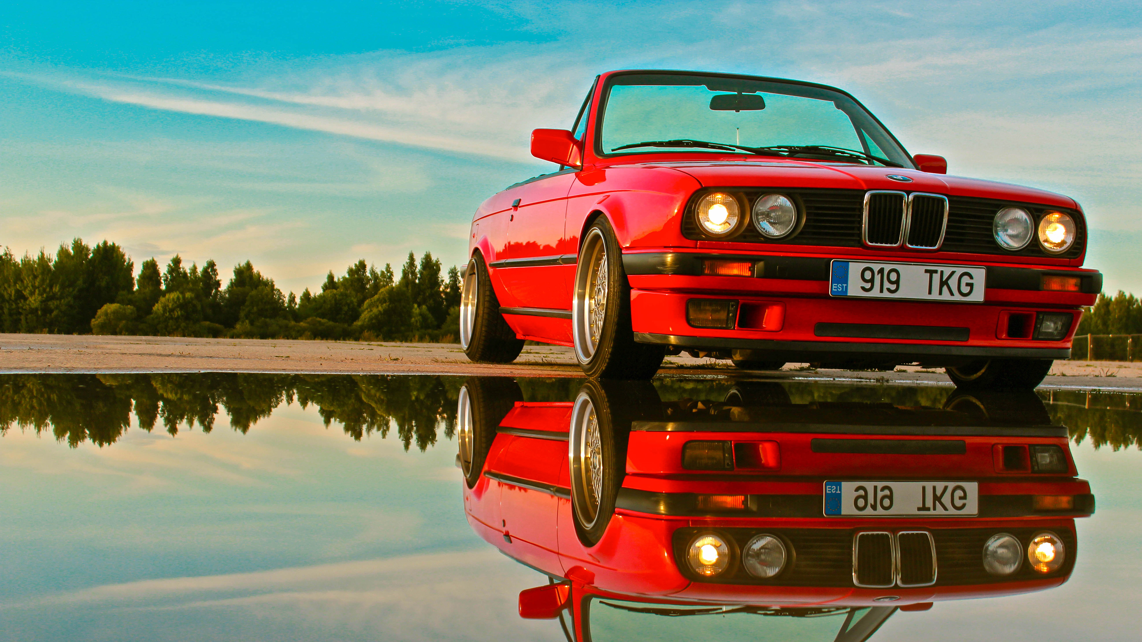 Red BMW E30 convertible parked near water with reflection, trees in background