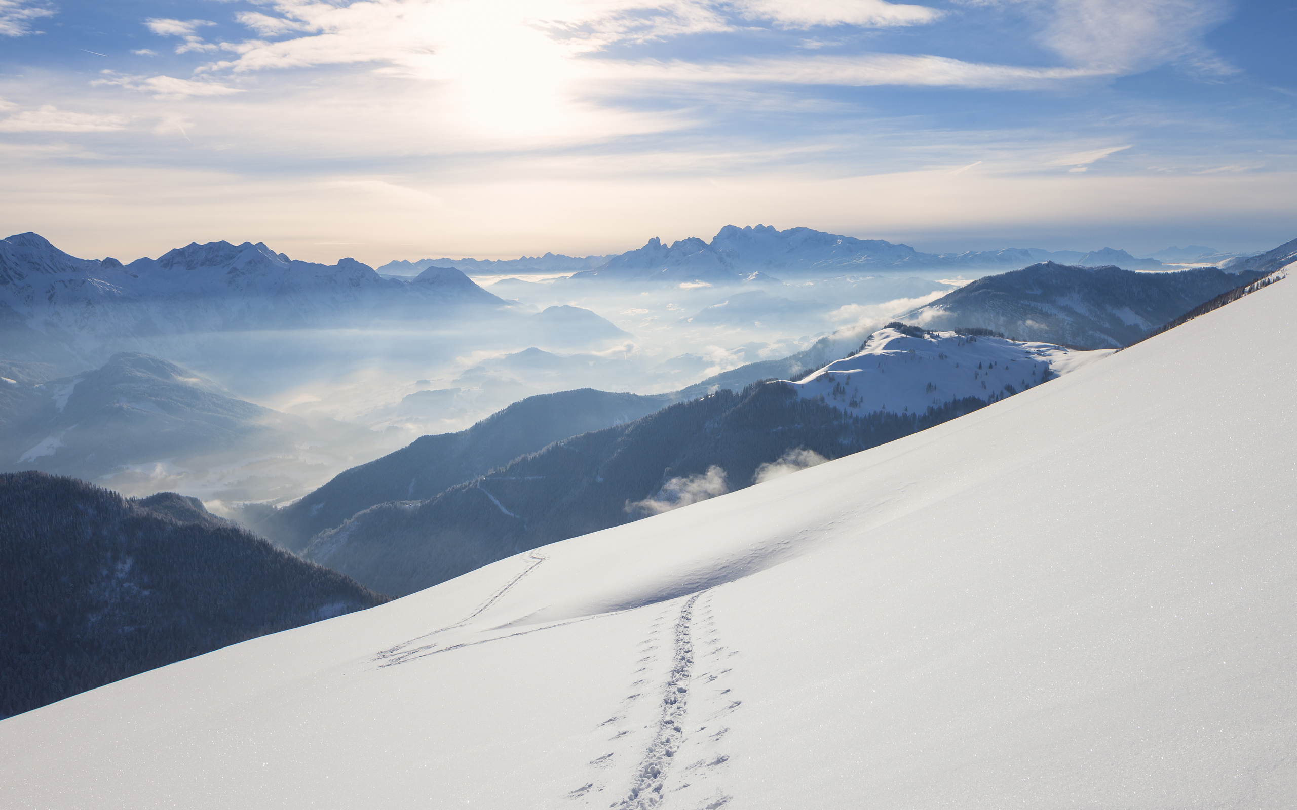 Snowy mountain slope with footprints leading down towards a misty valley and distant peaks