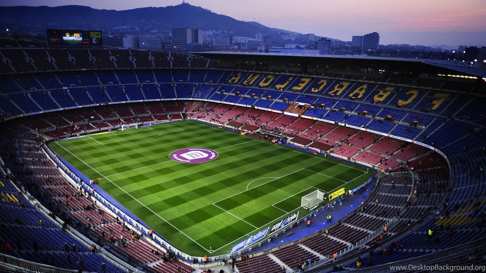 A panoramic view of Camp Nou stadium in Barcelona, Spain, during twilight. The vibrant green pitch is marked for a football match, and the stadium seats are partially filled with spectators. The stadium lights illuminate the scene, and the 'FC BARCELONA' lettering is clearly visible in the stands