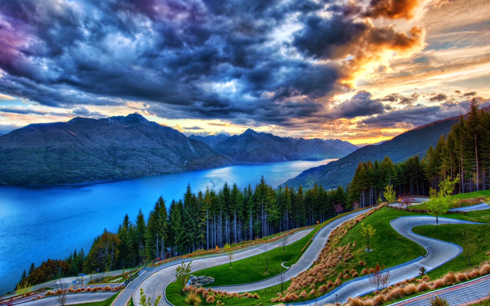 A breathtaking sunset view over a vibrant blue lake nestled amidst majestic mountains, with a winding road leading down through lush greenery