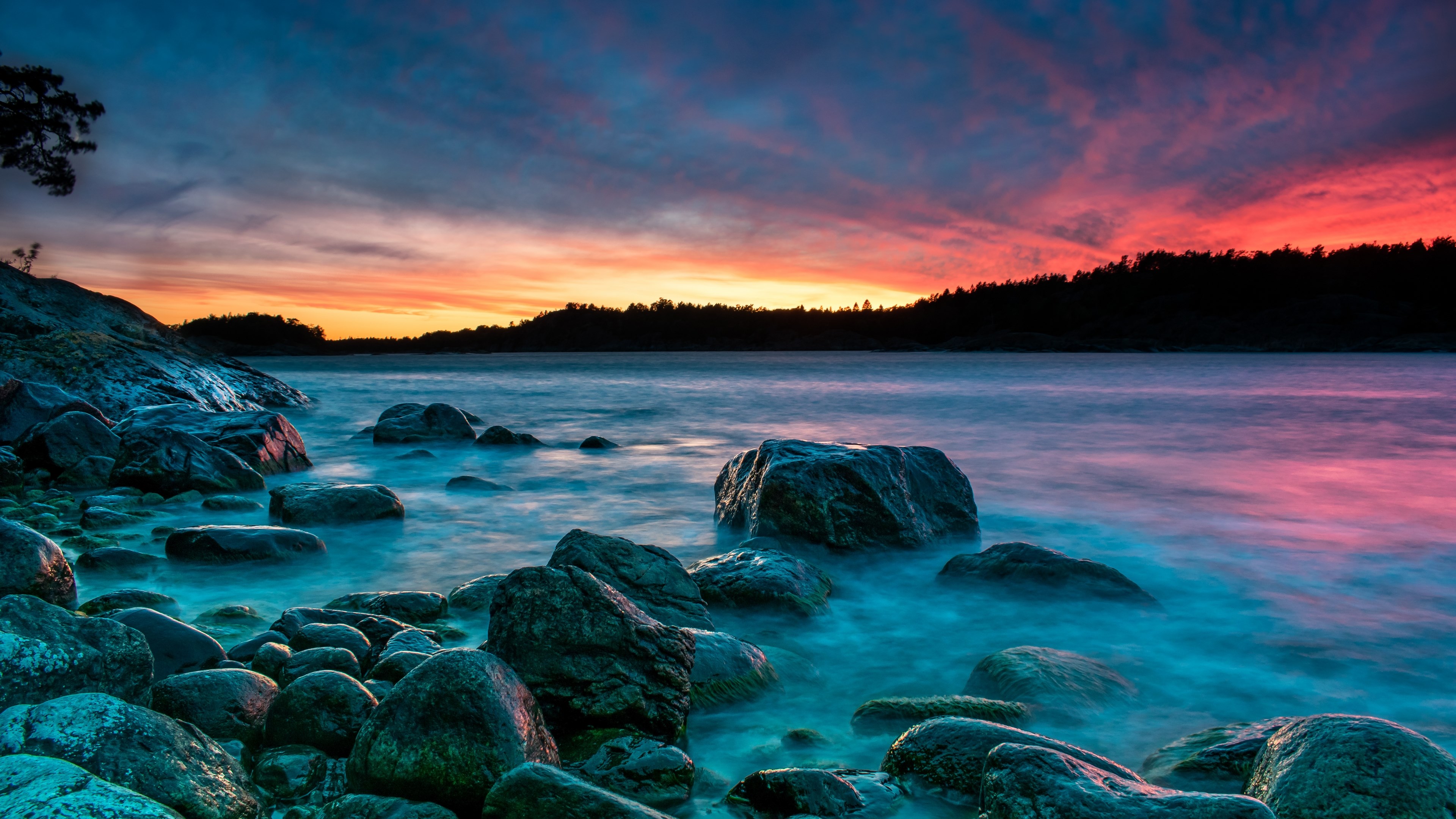 Stunning 4K wallpaper of a vibrant sunset over a rocky coastline. The smooth, colorful water contrasts beautifully with the dark silhouettes of the rocks and trees