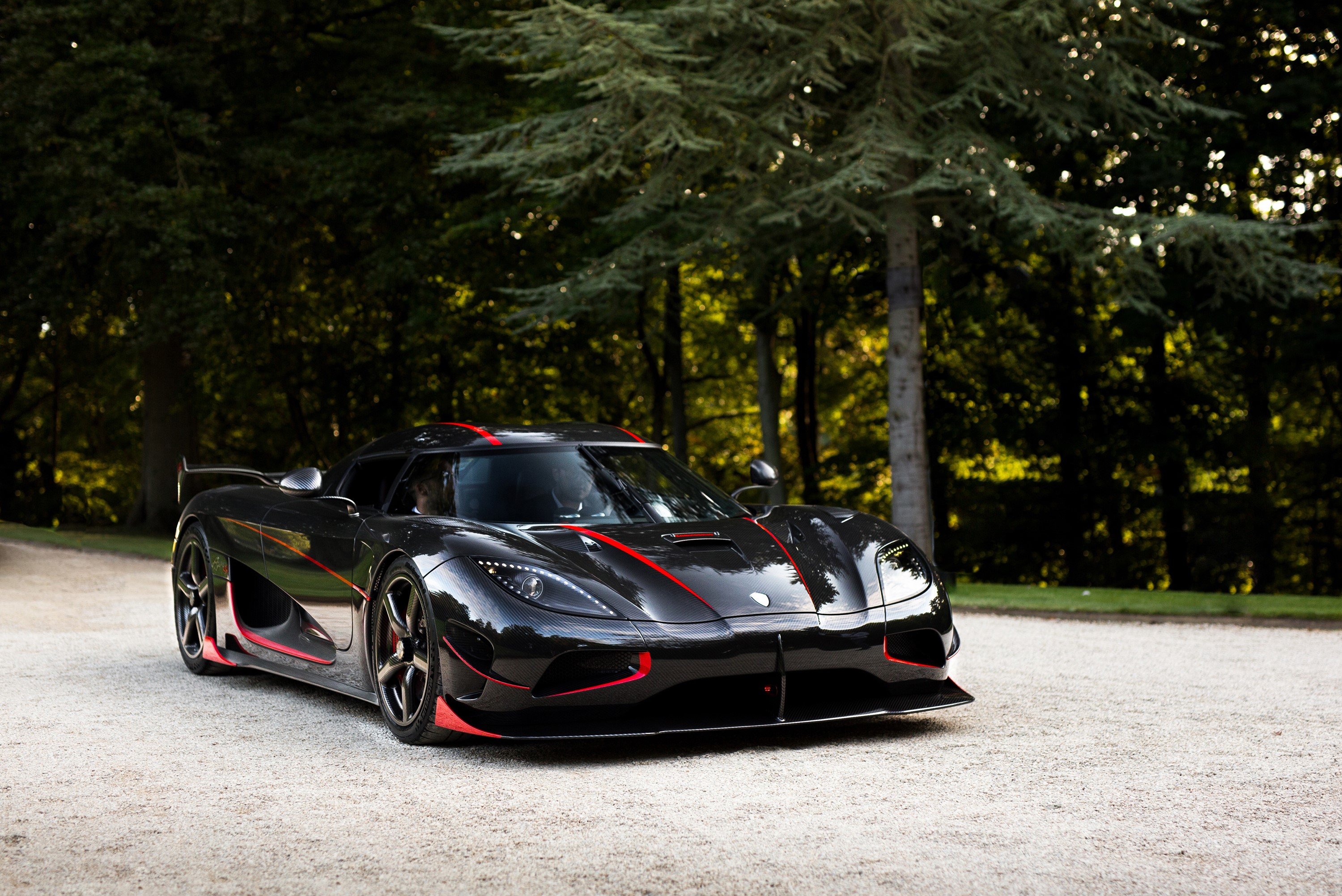 A sleek black Koenigsegg Regera with red accents parked on a gravel driveway, surrounded by lush green trees