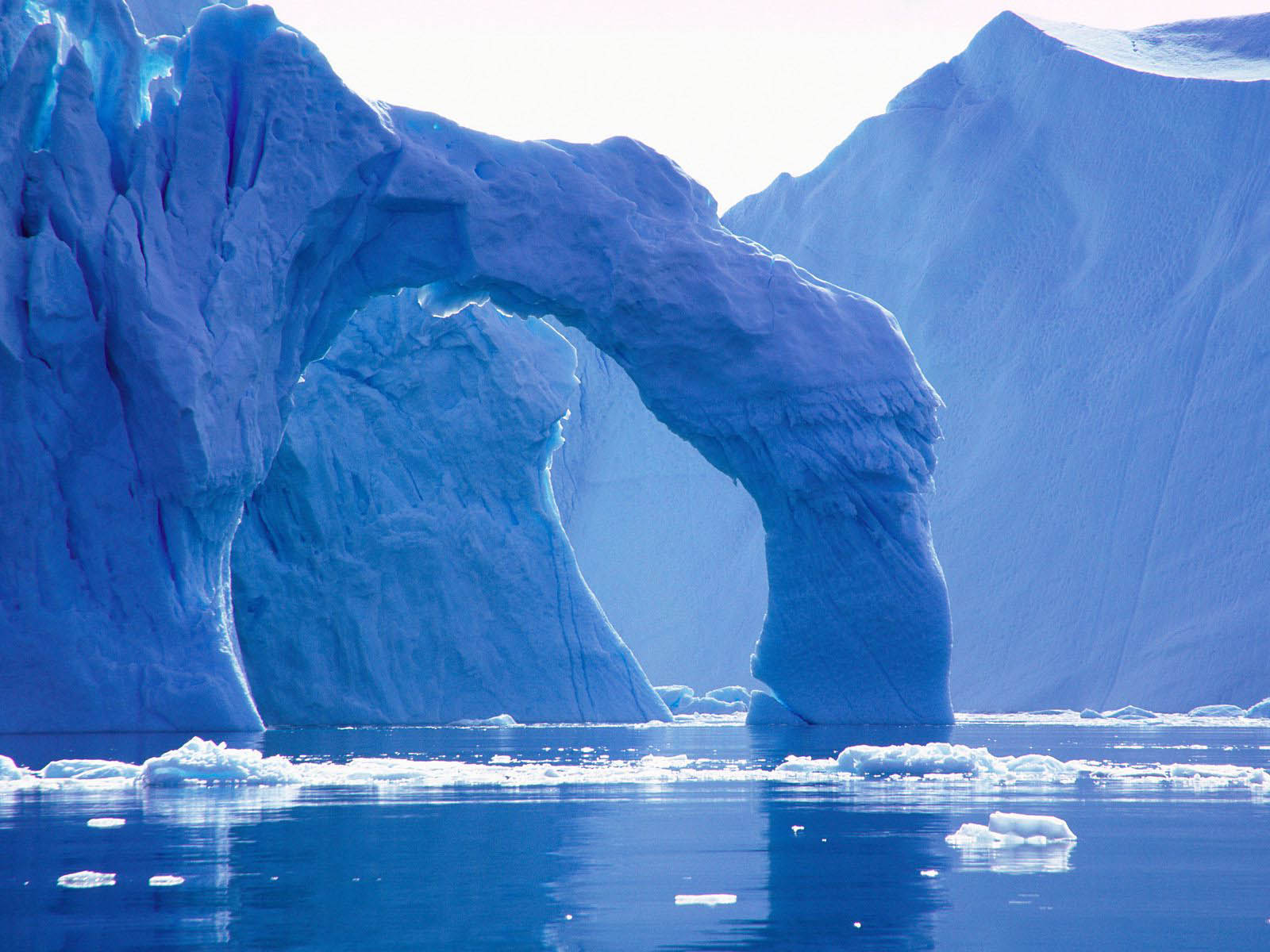 A breathtaking natural ice arch formed in a massive iceberg in the Arctic or Antarctic, with smaller ice floes dotting the serene blue water. The sunlight illuminates the icy structure, creating a stunning contrast with the deep blue hues of the sea and ice