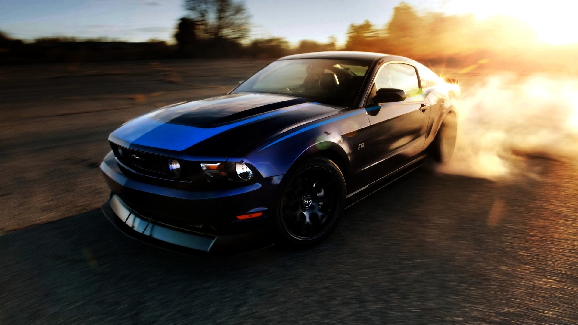 A dark blue Ford Mustang drifting on a paved road at sunset, with smoke billowing from the tires