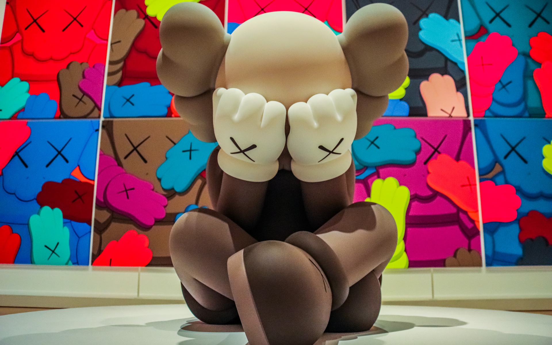 A KAWS Companion figure sits with its hands covering its eyes, in front of a vibrant, colorful artwork featuring similar figures. This piece showcases KAWS' signature style of playful yet melancholic characters