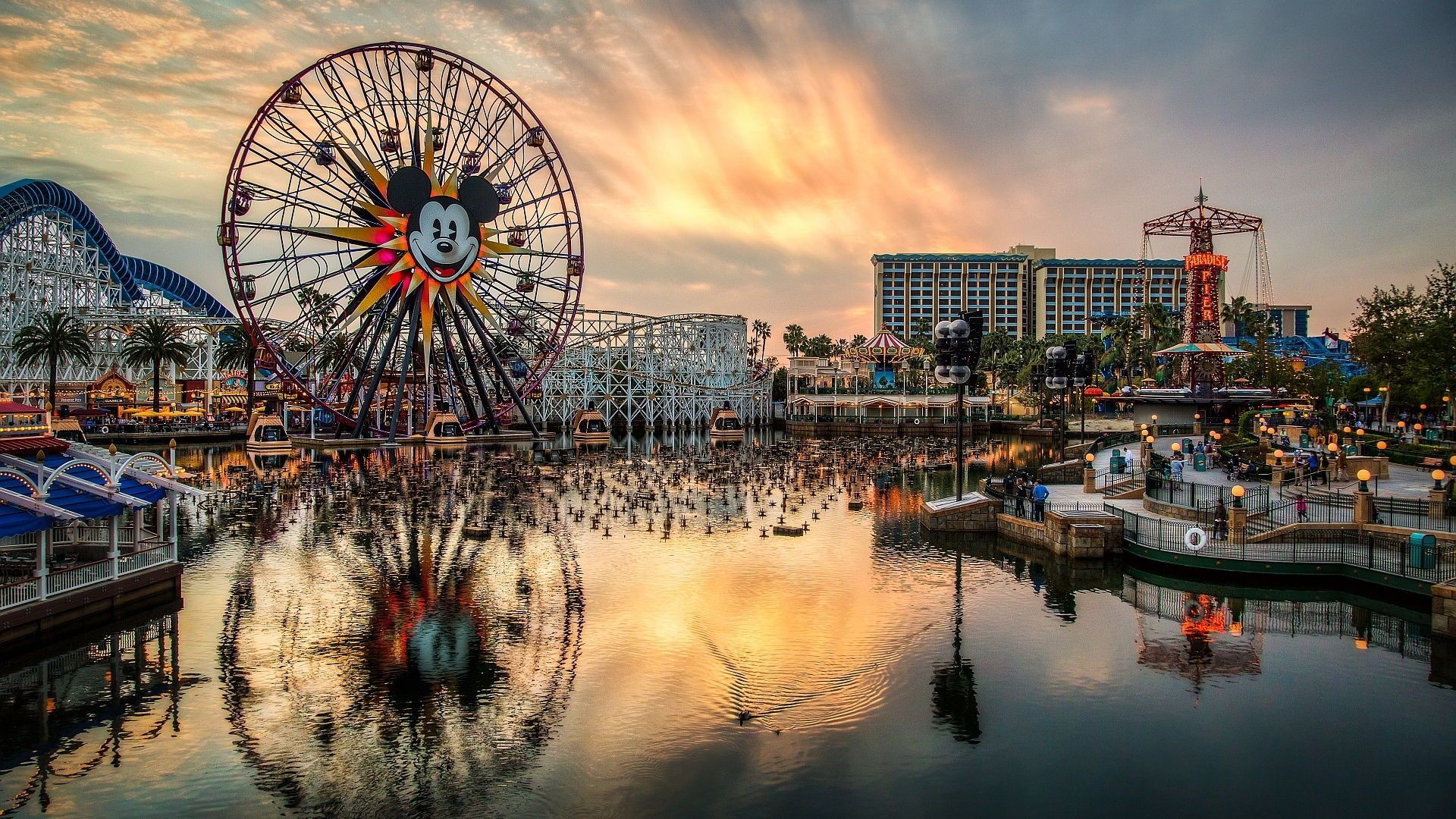 A stunning sunset over Paradise Pier at Disney California Adventure Park, featuring Mickey's Fun Wheel and the California Screamin' roller coaster reflected in Paradise Bay