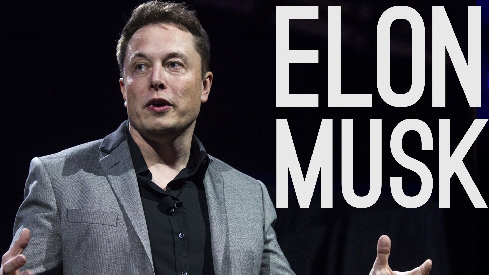 Elon Musk speaking at an event. He is wearing a gray suit jacket and a black shirt