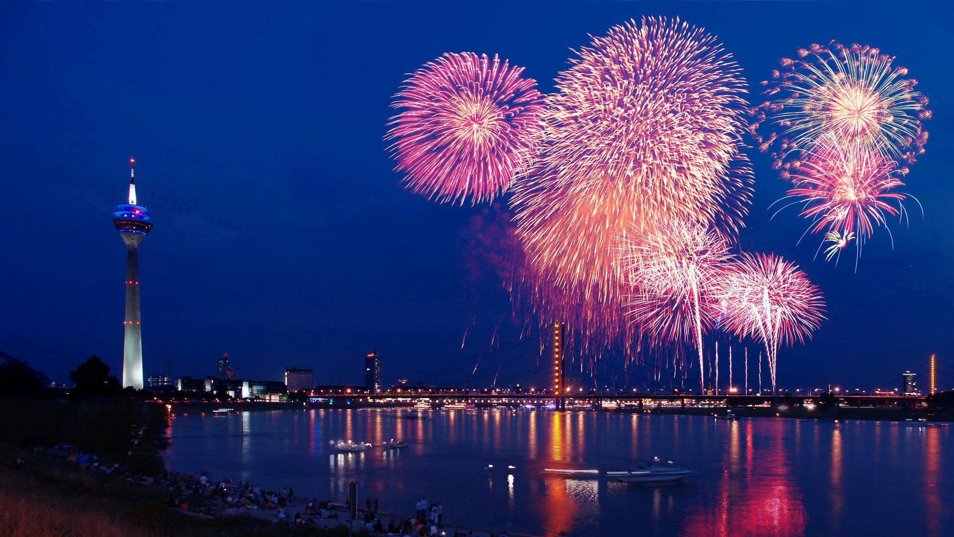 Fireworks light up the night sky over a city skyline with a prominent tower, celebrating New Year's Eve