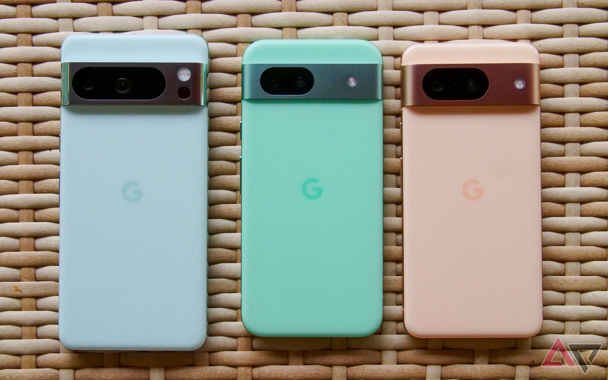 Three Google Pixel 7a phones in Snow, Sea, and Coral color variants displayed on a woven surface