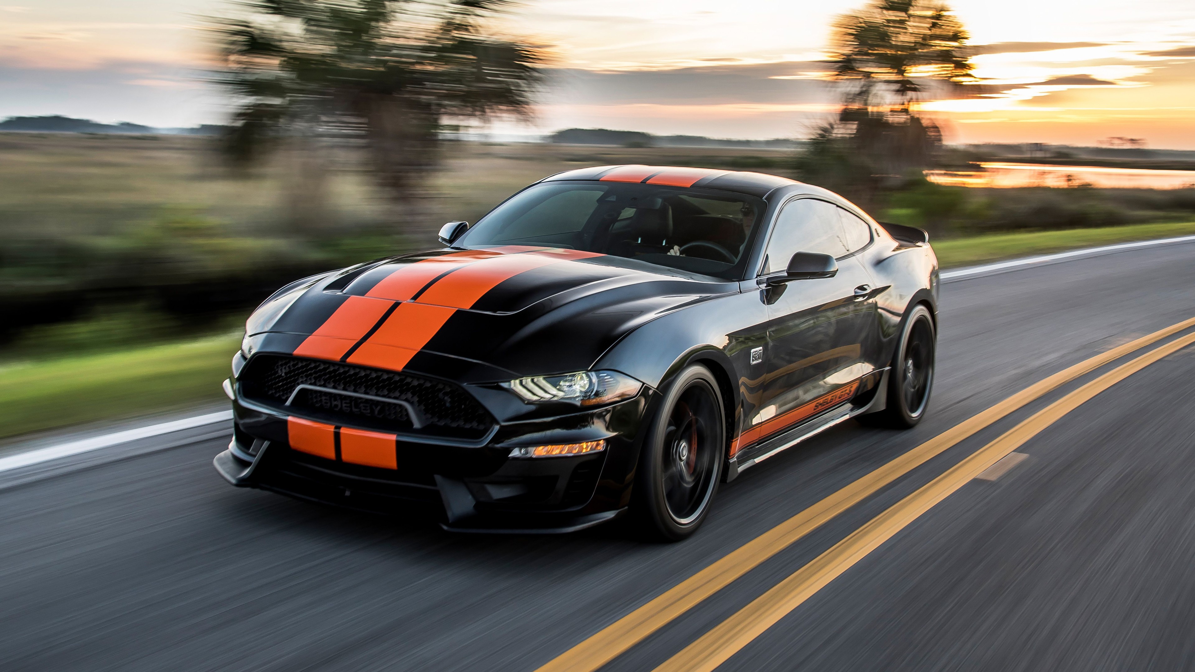 A black Ford Mustang Shelby GT500 with orange racing stripes speeding down a scenic highway at sunset