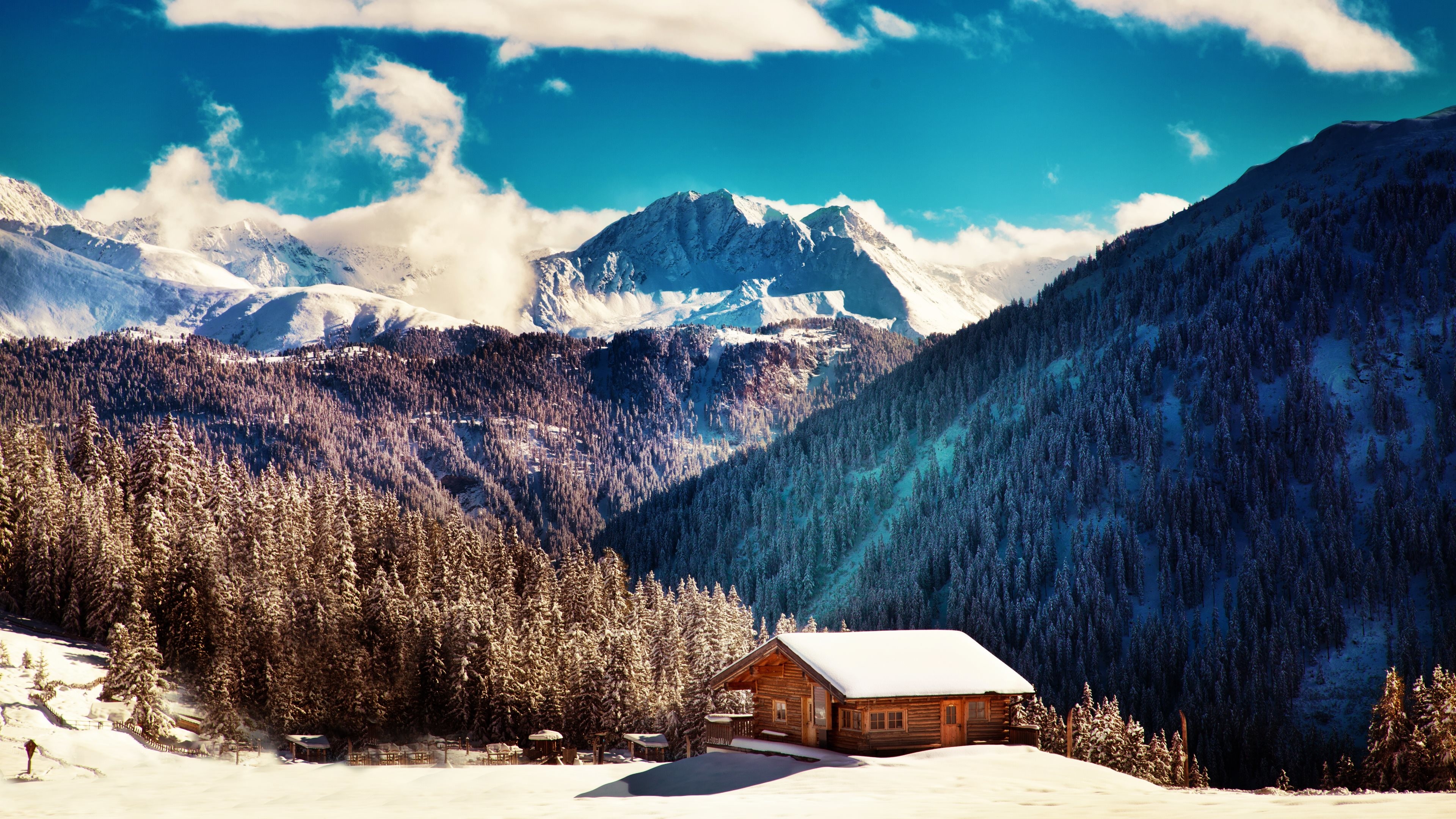 A cozy wooden cabin nestled in a snowy valley amidst snow-covered pine trees and majestic mountains under a vibrant blue sky