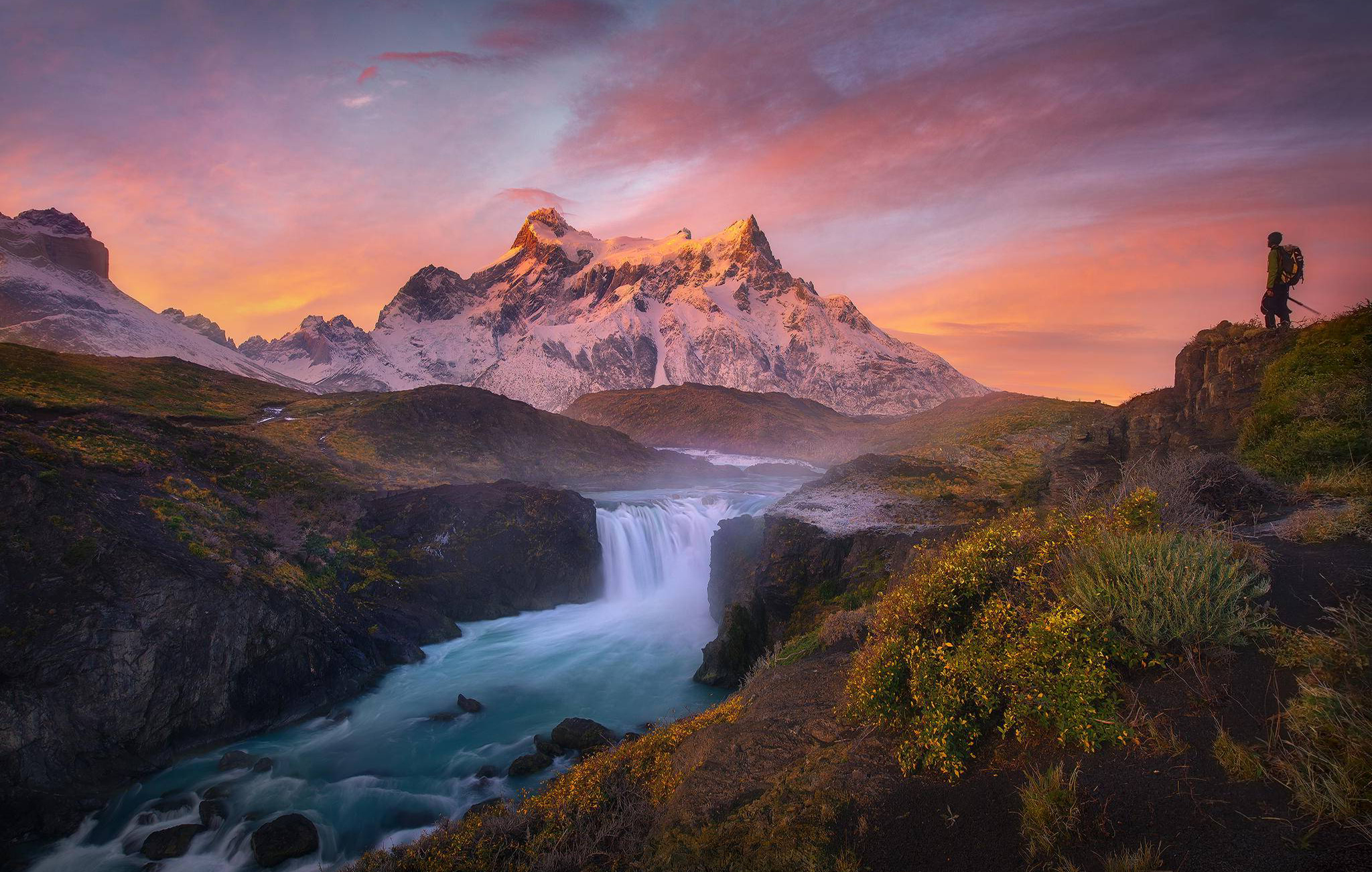 A breathtaking landscape photograph of a waterfall cascading into a turquoise river, with snow-capped mountains in the background and a hiker silhouetted against the vibrant sunset sky