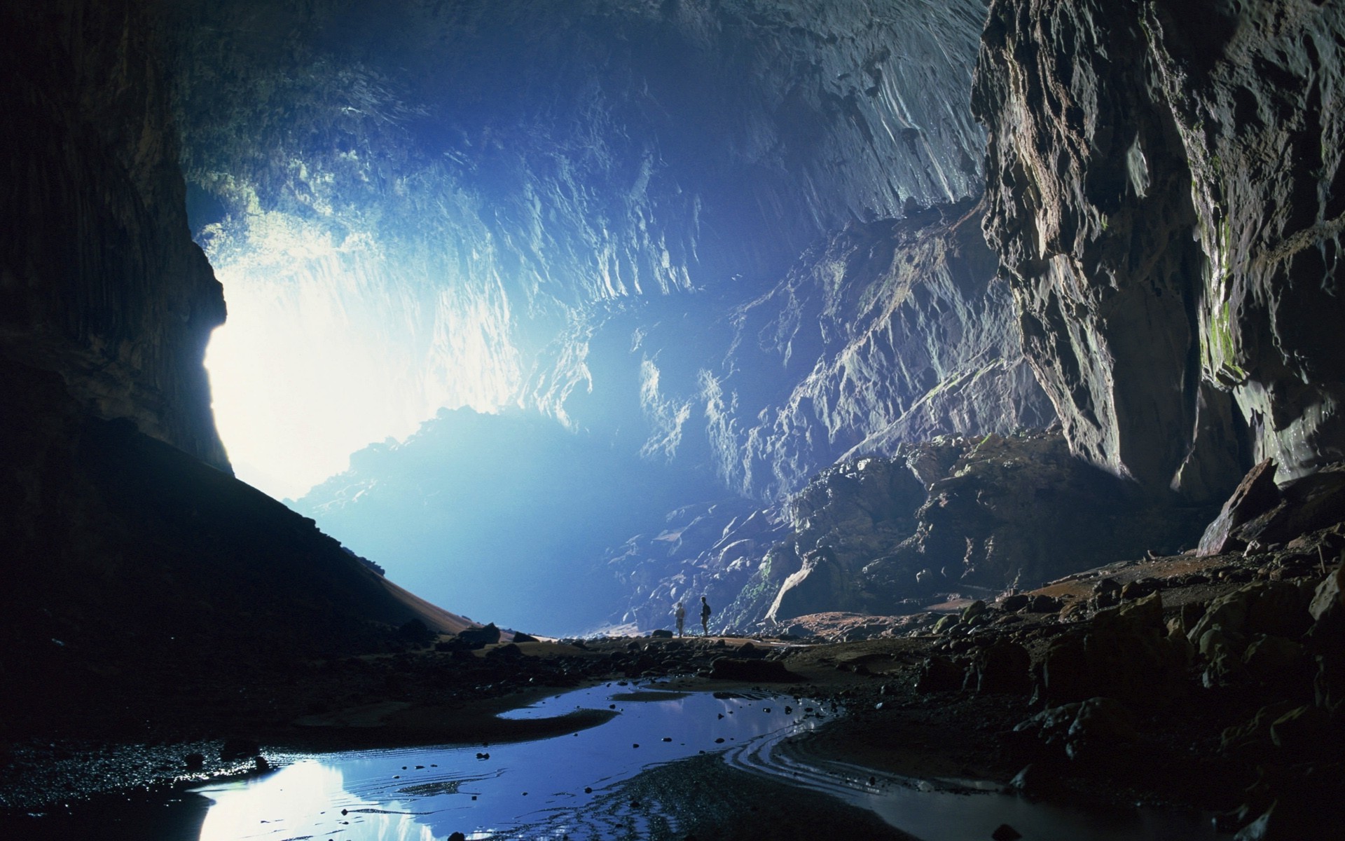 Sunlight streams into a vast cave, illuminating two small figures exploring its depths
