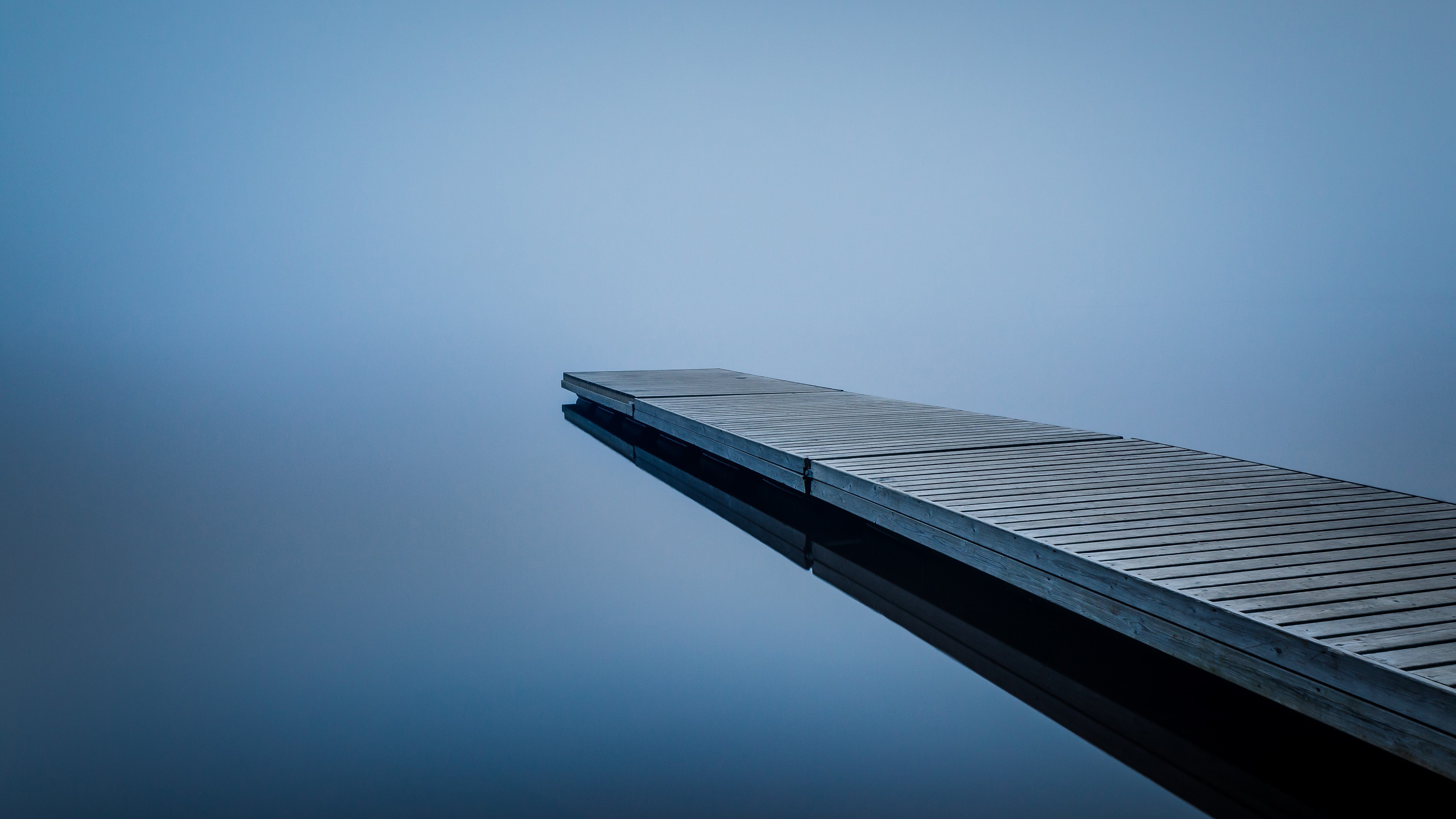 A serene 4K wallpaper featuring a wooden dock extending into a calm, misty lake. The soft blue hues of the water and sky create a peaceful and minimalist scene