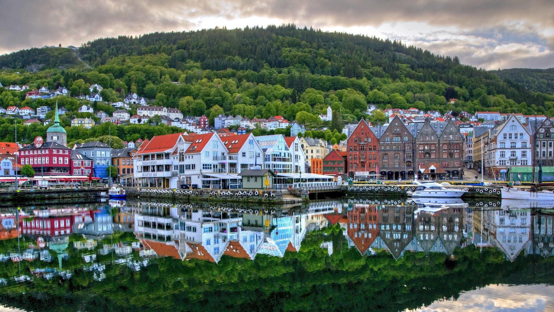 Colorful houses reflected in the calm waters of a harbor in a cozy city, with lush green hills rising in the background