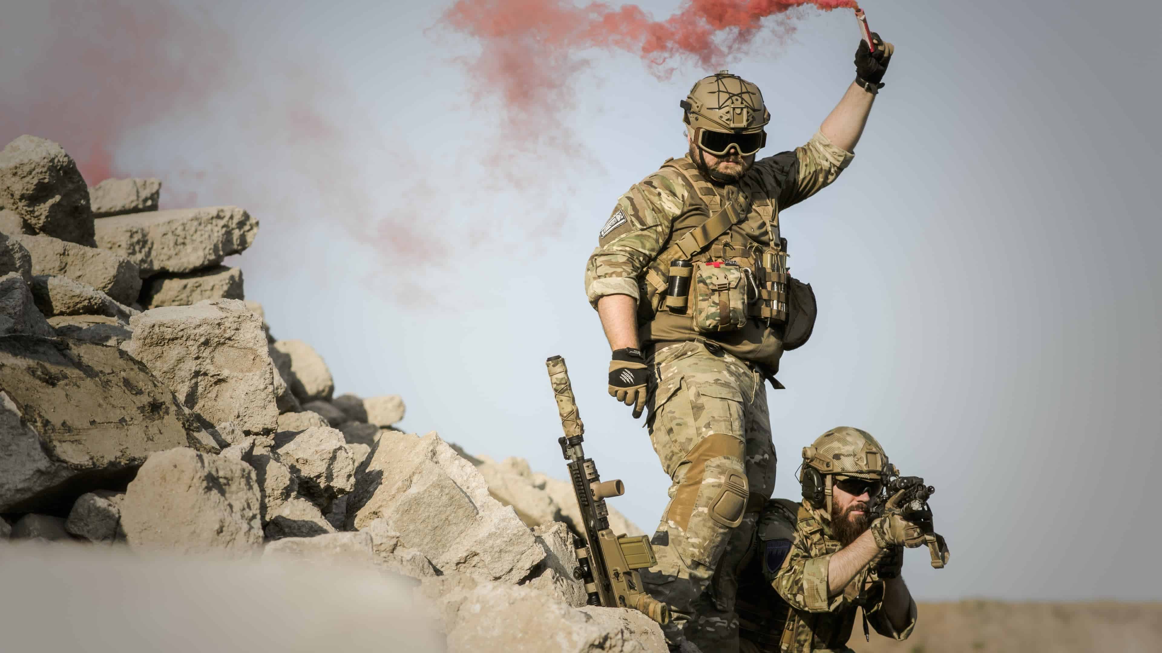 Two soldiers in camouflage gear, one holding a red smoke flare and the other aiming a rifle, stand amidst a pile of rubble