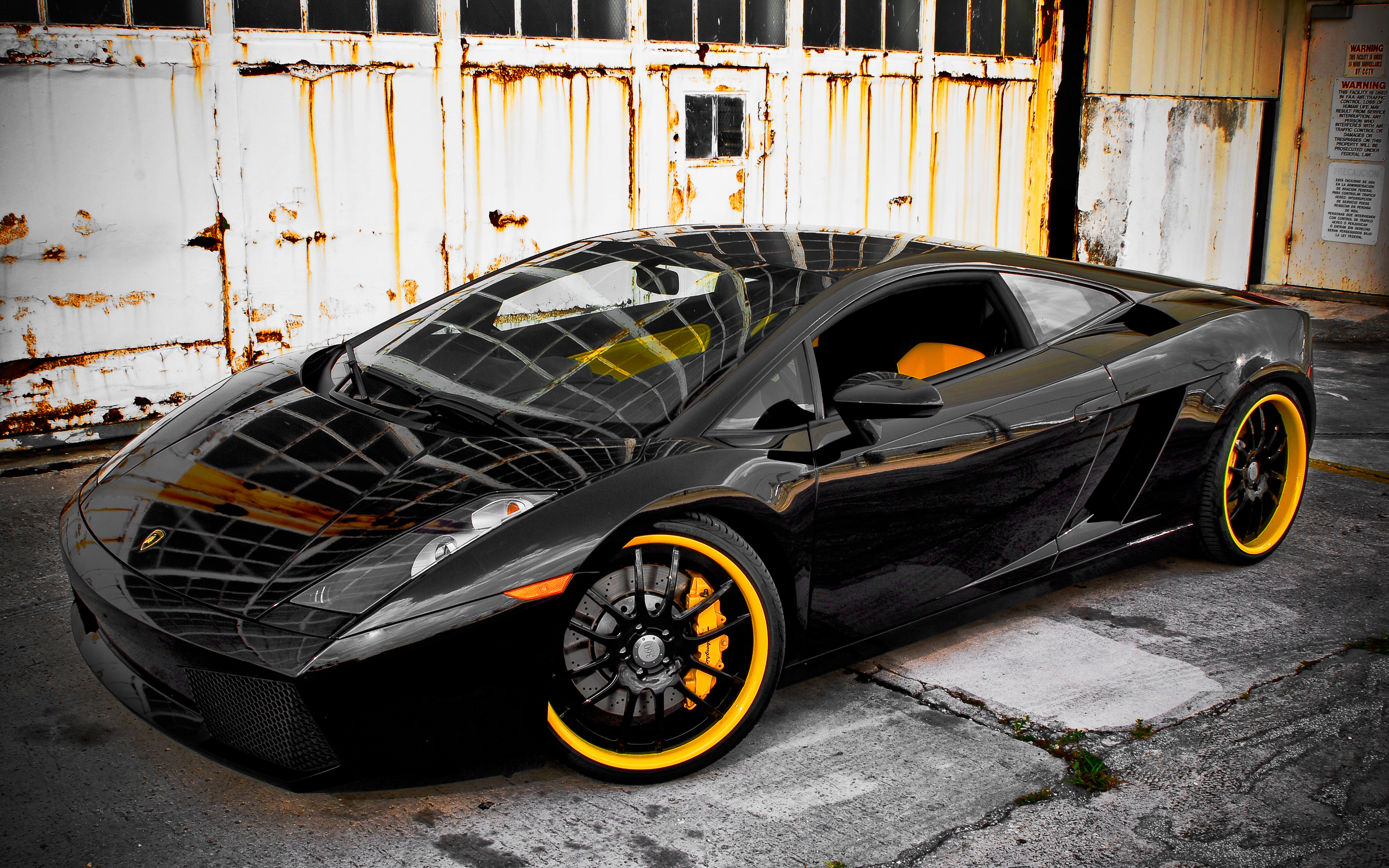 A sleek black Lamborghini Gallardo with vibrant yellow rims parked in front of a weathered industrial backdrop