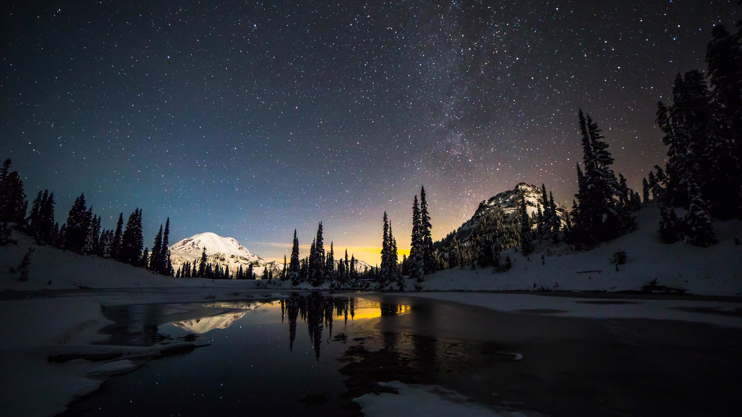A breathtaking 5K wallpaper showcasing a snow-covered mountain range at night, reflected serenely in a partially frozen lake, under a star-studded sky