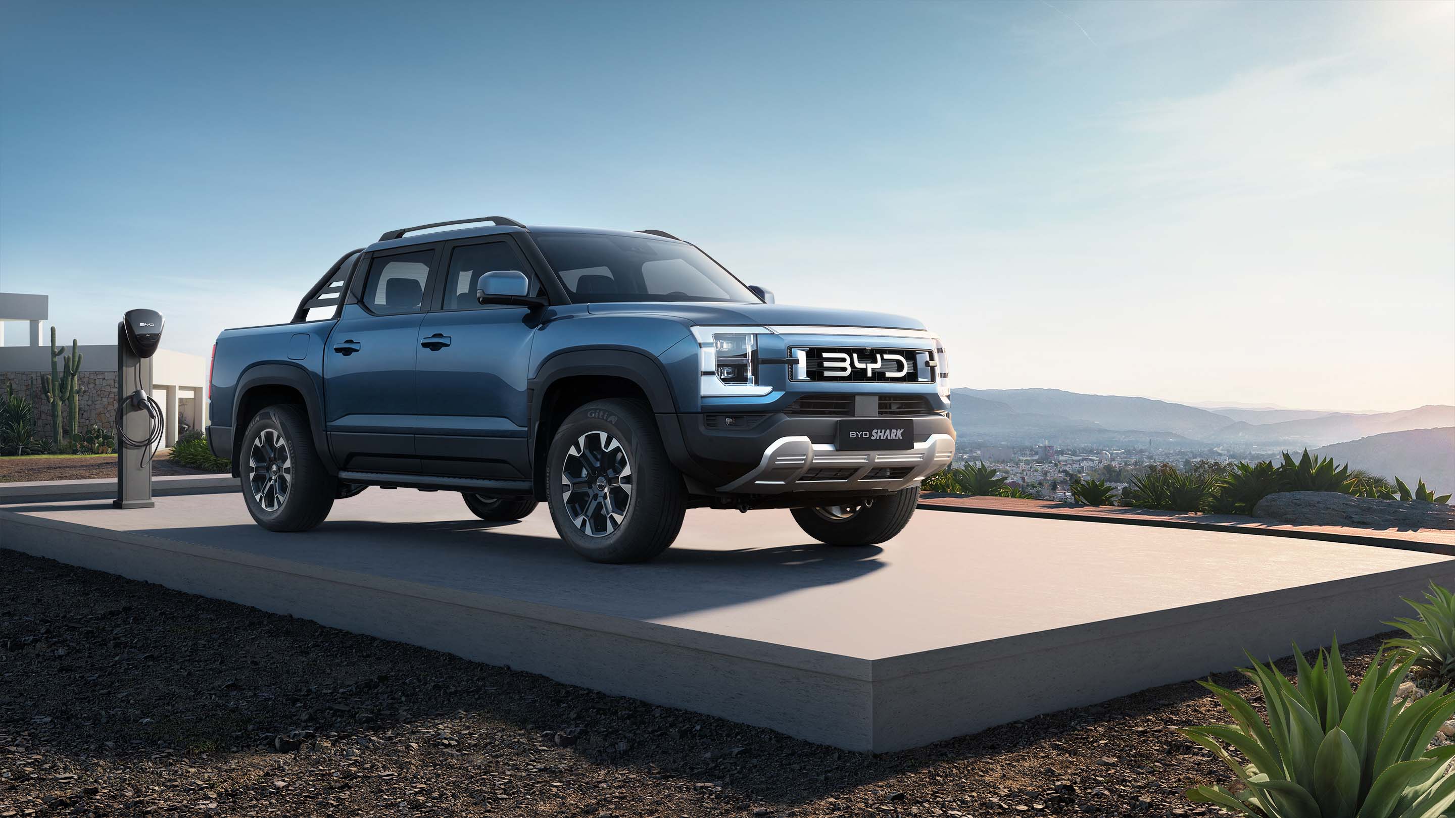 A blue BYD Shark electric pickup truck parked at a charging station overlooking a scenic valley