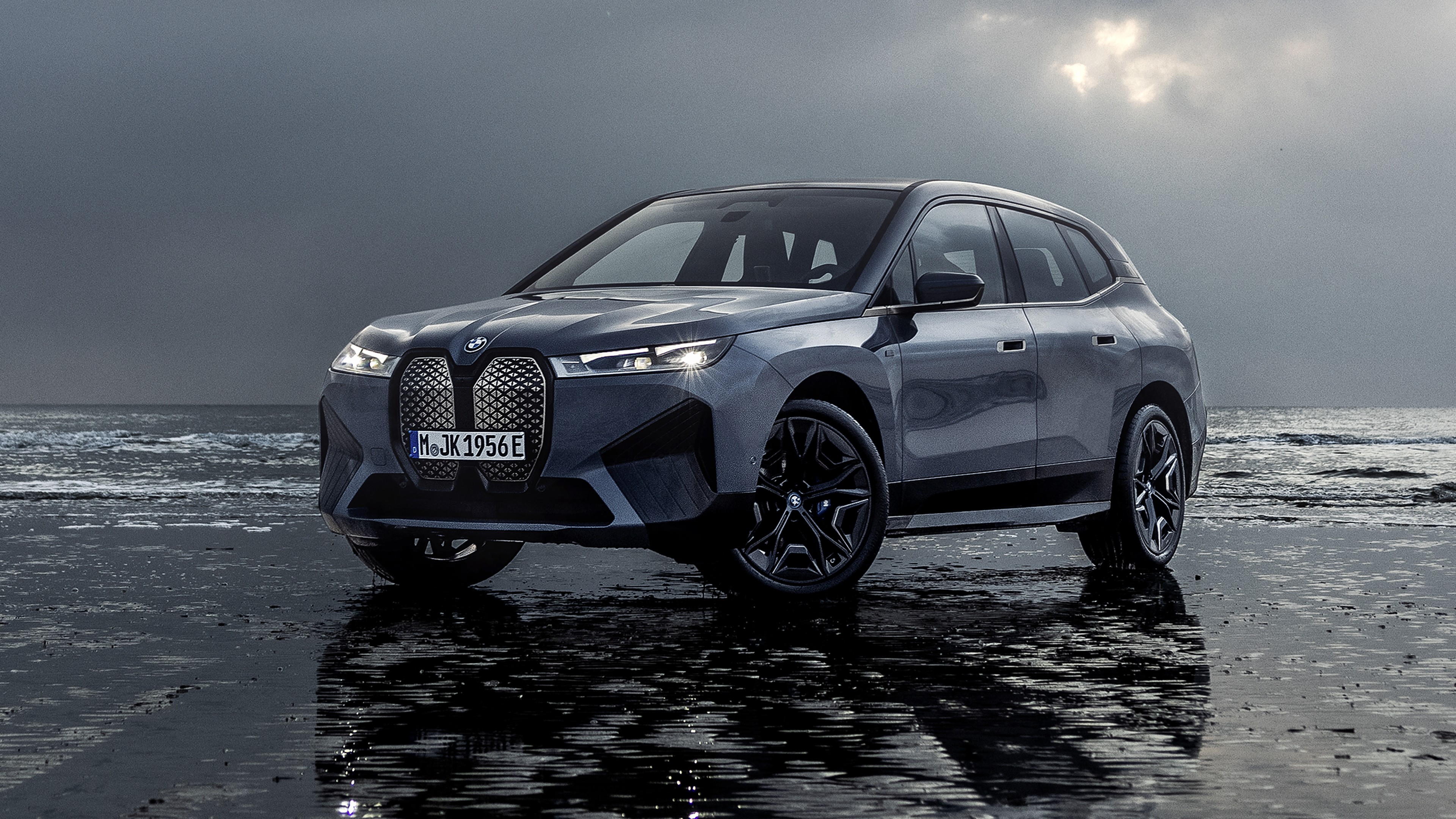 A sleek, gray BMW iX electric SUV parked on a wet, reflective beach with a dramatic sky overhead