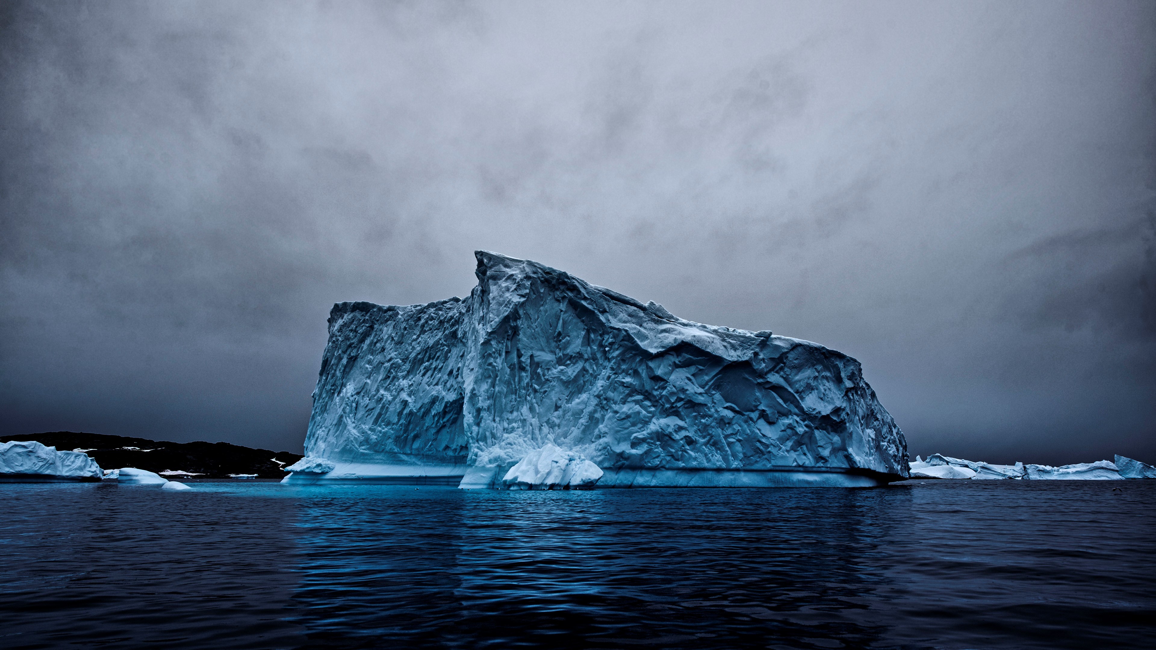 A majestic iceberg floats in dark blue water under a stormy gray sky. The iceberg's textured surface and imposing size create a dramatic scene