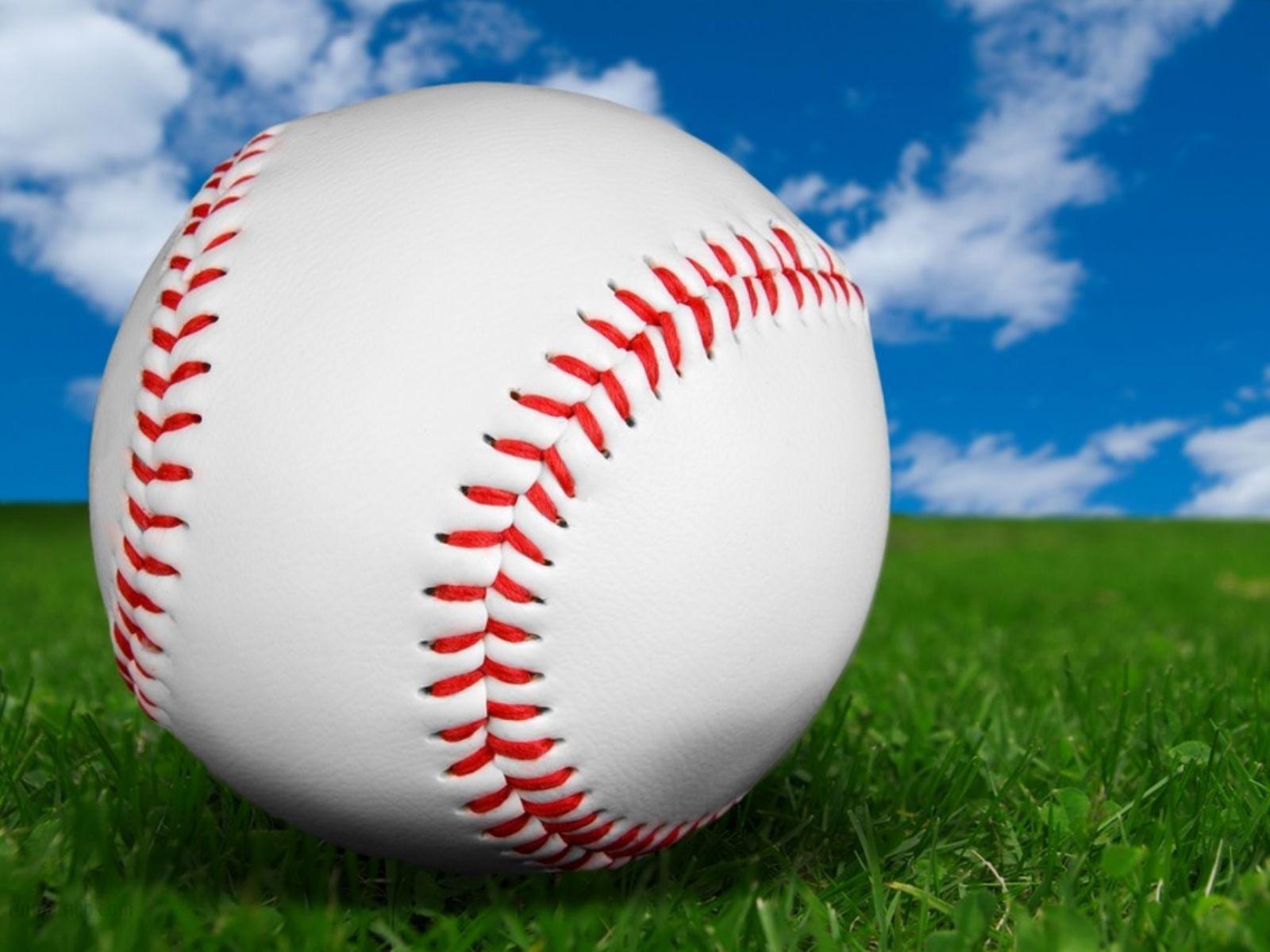 A close-up of a baseball with red stitching, resting on a vibrant green grass field against a backdrop of a clear blue sky dotted with fluffy white clouds
