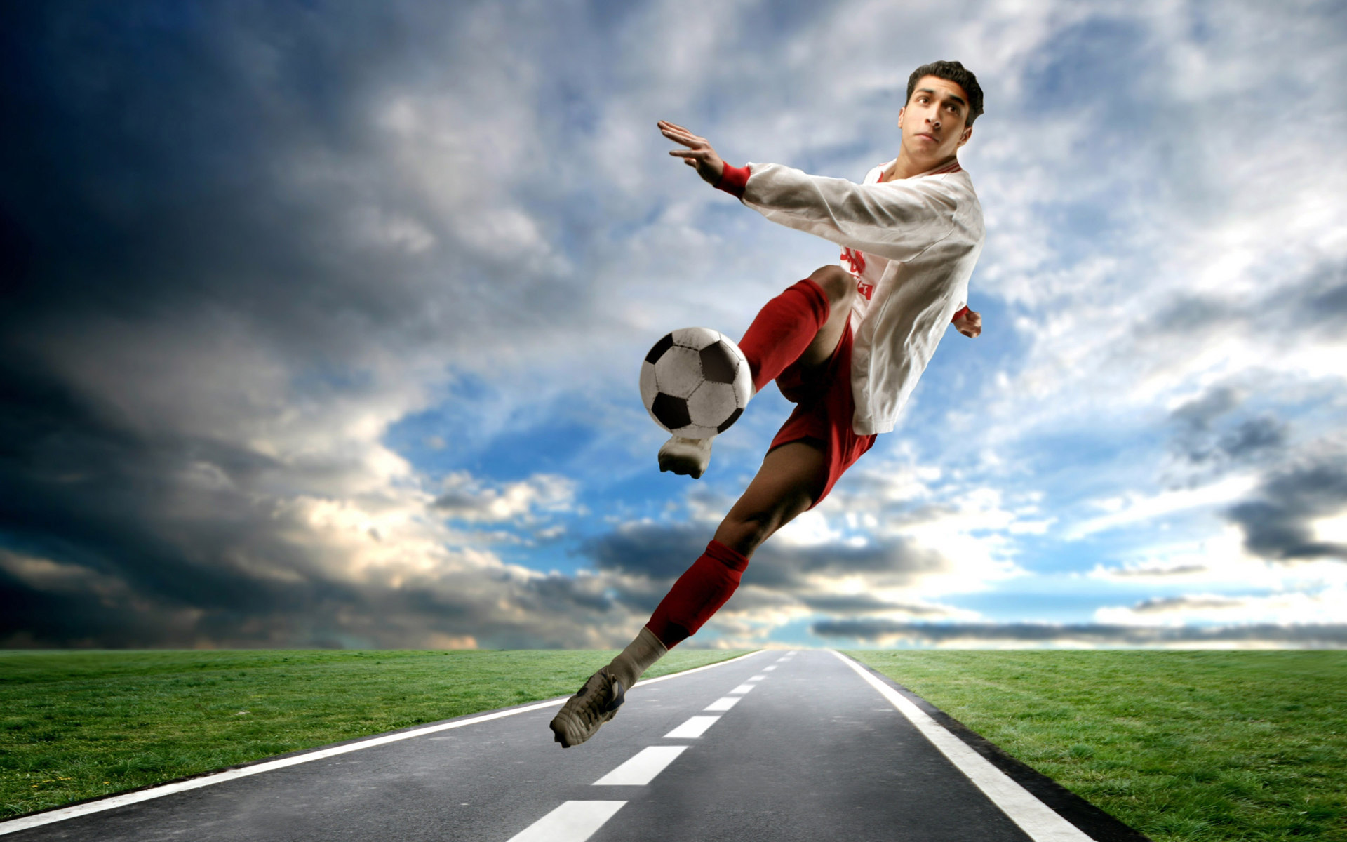 A soccer player dynamically kicks the ball mid-air above a paved road, set against a dramatic sky and green fields