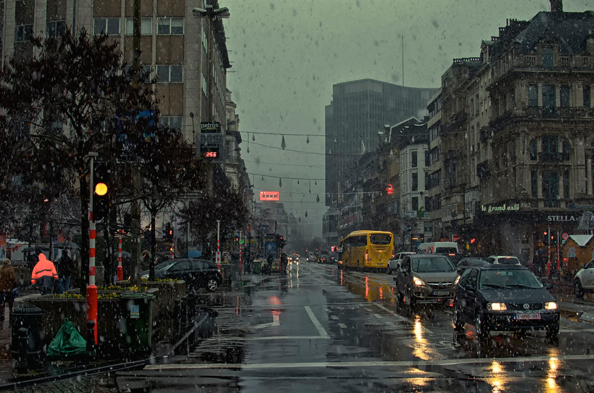 A rainy city street scene at dusk, with cars and buses driving through the wet streets. The streetlights reflect in the puddles, creating a moody and atmospheric image
