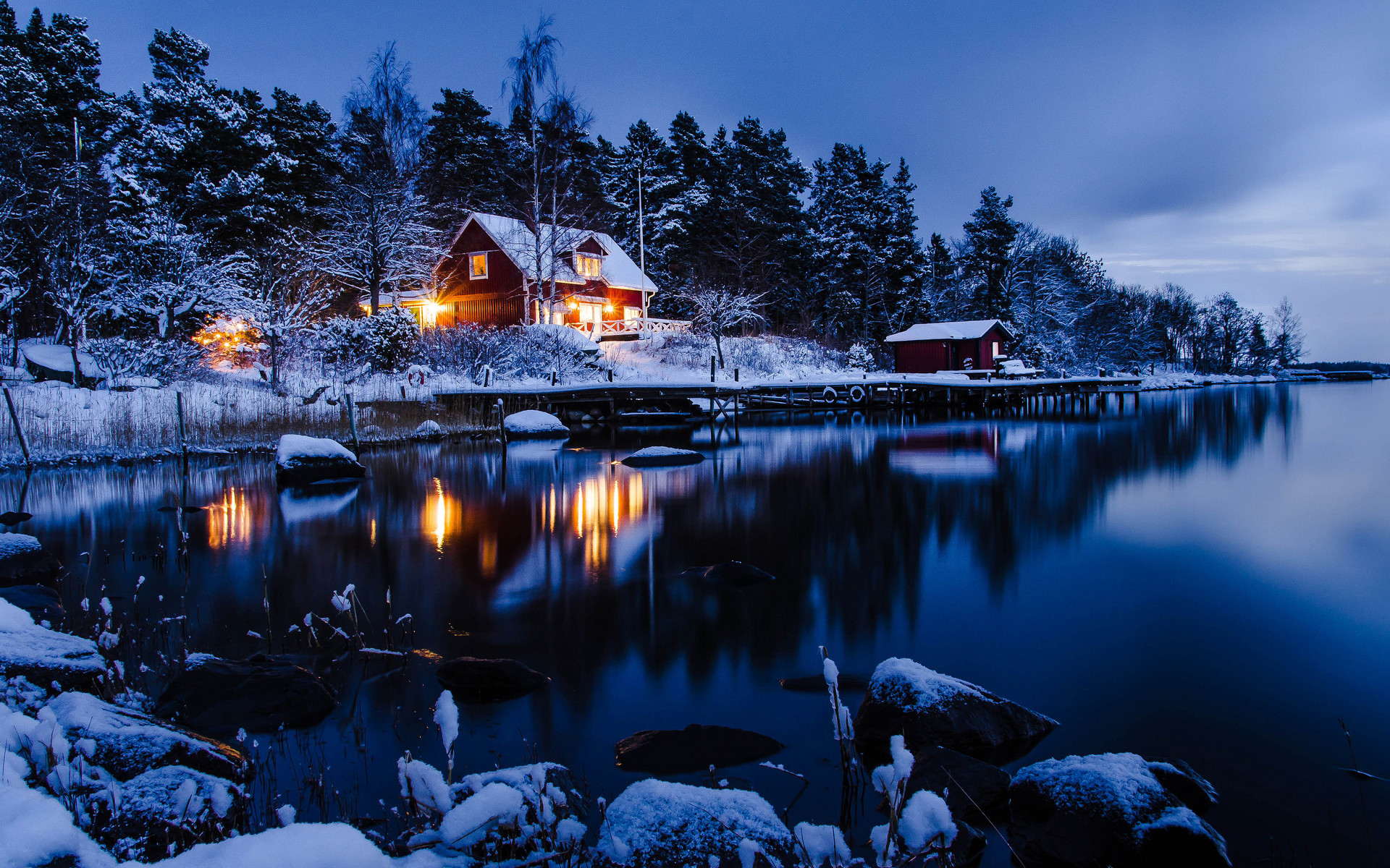 A cozy red house glows warmly against the twilight sky, nestled among snow-covered trees on the edge of a tranquil lake. A small, snow-dusted dock extends into the still water, reflecting the lights of the house and the surrounding winter landscape