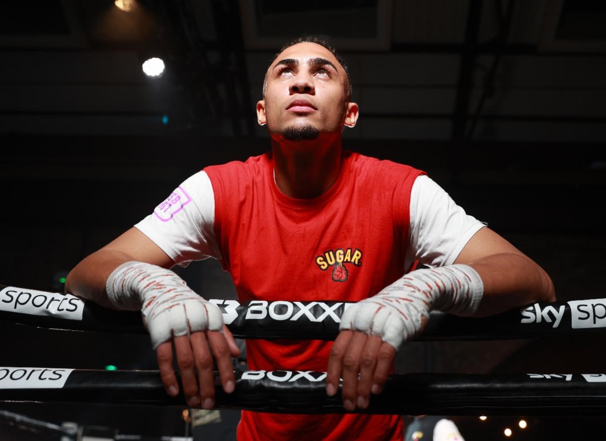 Benjamin Whittaker, a rising star in boxing, looks upwards in contemplation, leaning against the ropes of a boxing ring. His intense gaze and the red and white of his training attire convey a sense of determination and focus. This image captures a moment of reflection and readiness before a match