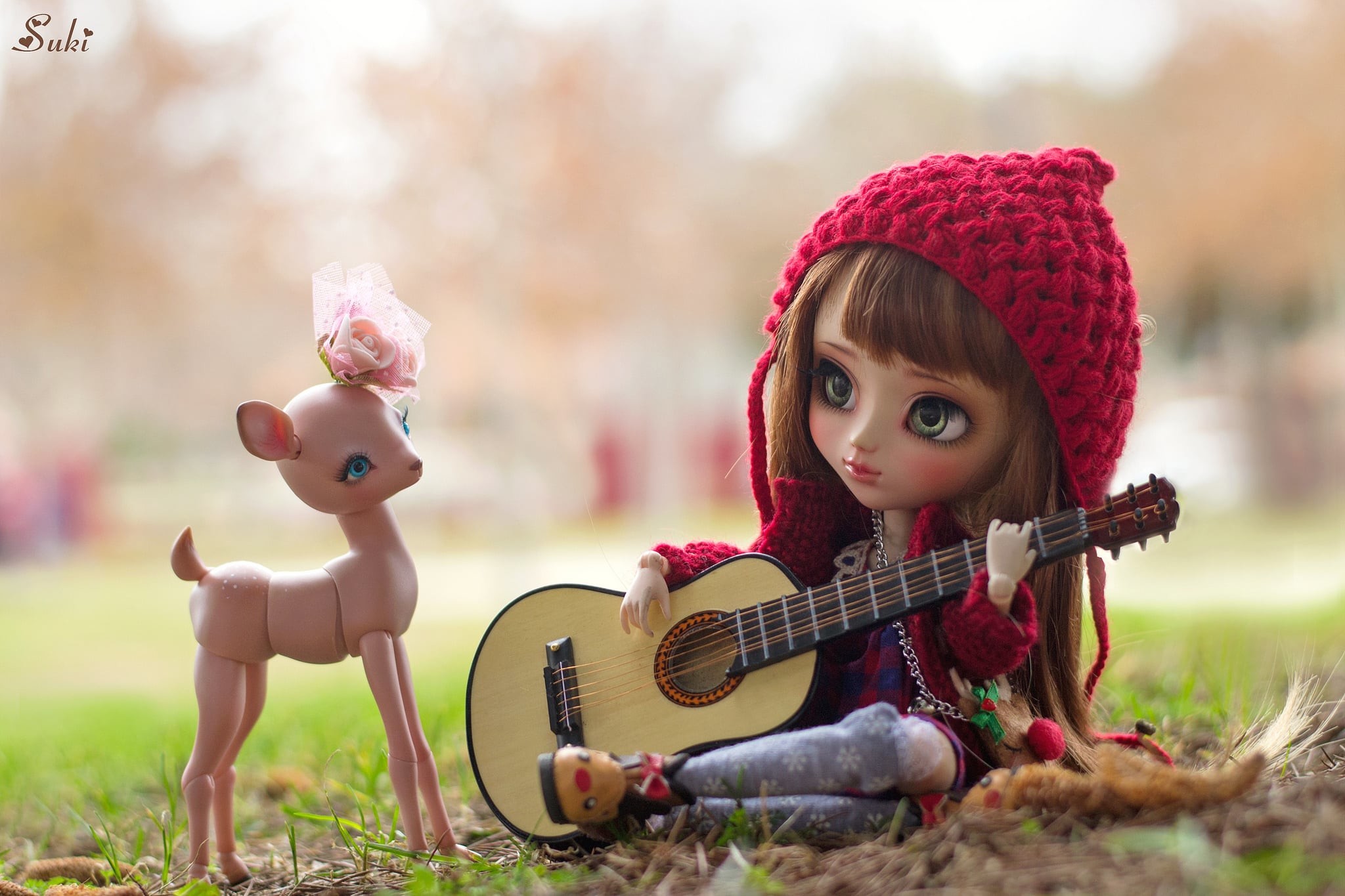 A doll with a red crocheted hood plays a miniature acoustic guitar for a small deer figurine in a grassy outdoor setting
