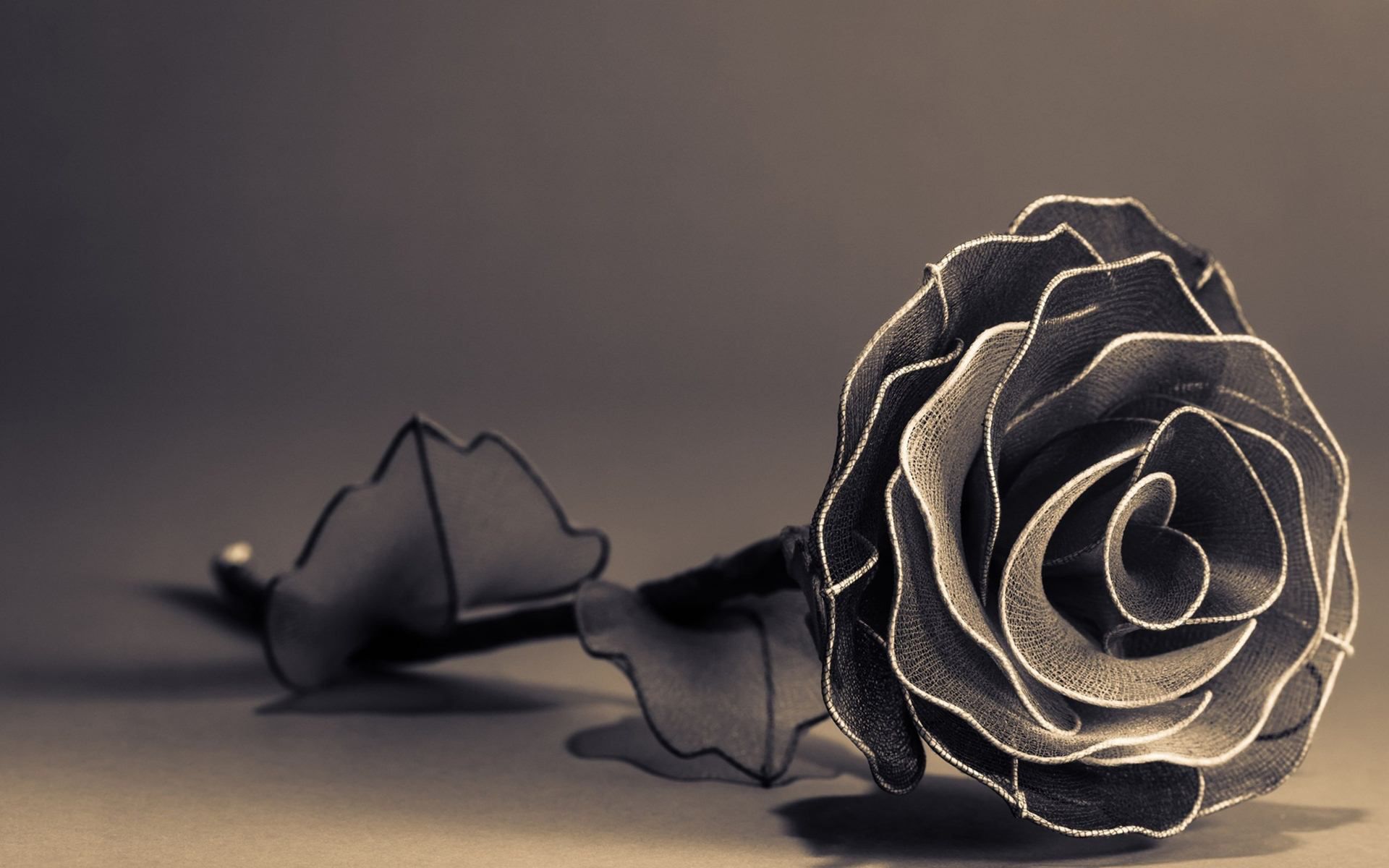 A monochromatic close-up of a handcrafted rose, meticulously formed from dark fabric and metallic threads. The delicate texture of the fabric and the subtle sheen of the threads create a captivating contrast, highlighting the intricate details of the rose's petals. The dark background enhances the rose's ethereal beauty, giving it an air of timeless elegance and artistic expression