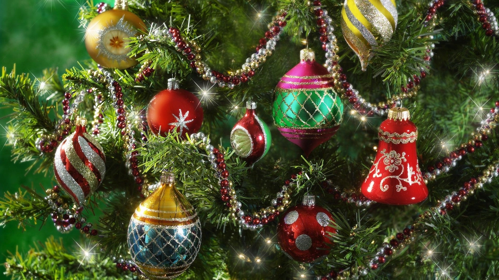 A close-up view of a Christmas tree adorned with colorful ornaments, including round baubles in various colors, a bell-shaped ornament, and a spiral-shaped ornament. A beaded garland winds through the branches, adding to the festive decorations