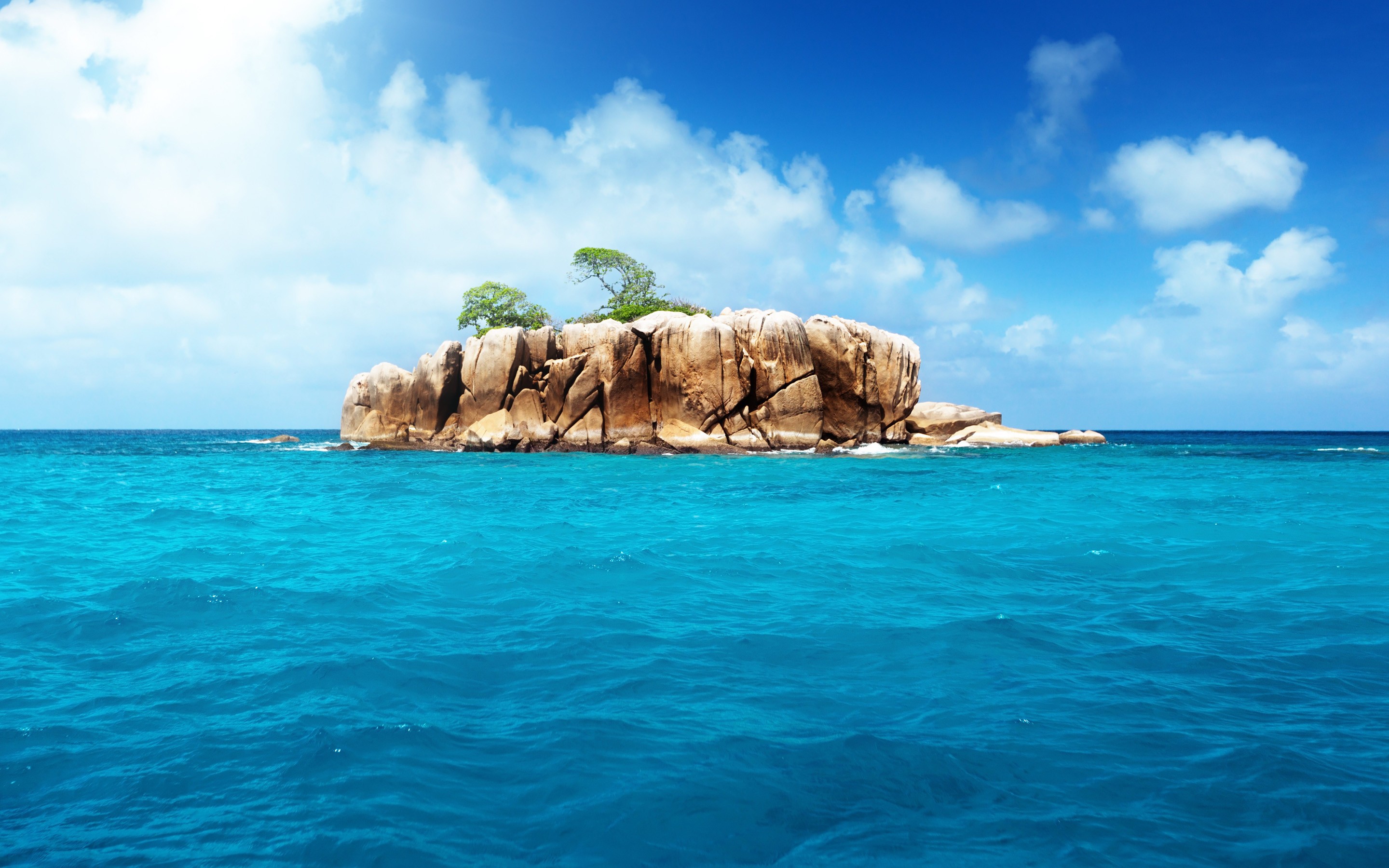 A small, rocky island with a few trees sits in the midst of a beautiful turquoise ocean under a bright blue sky with scattered clouds. This serene image evokes a sense of tranquility and escape
