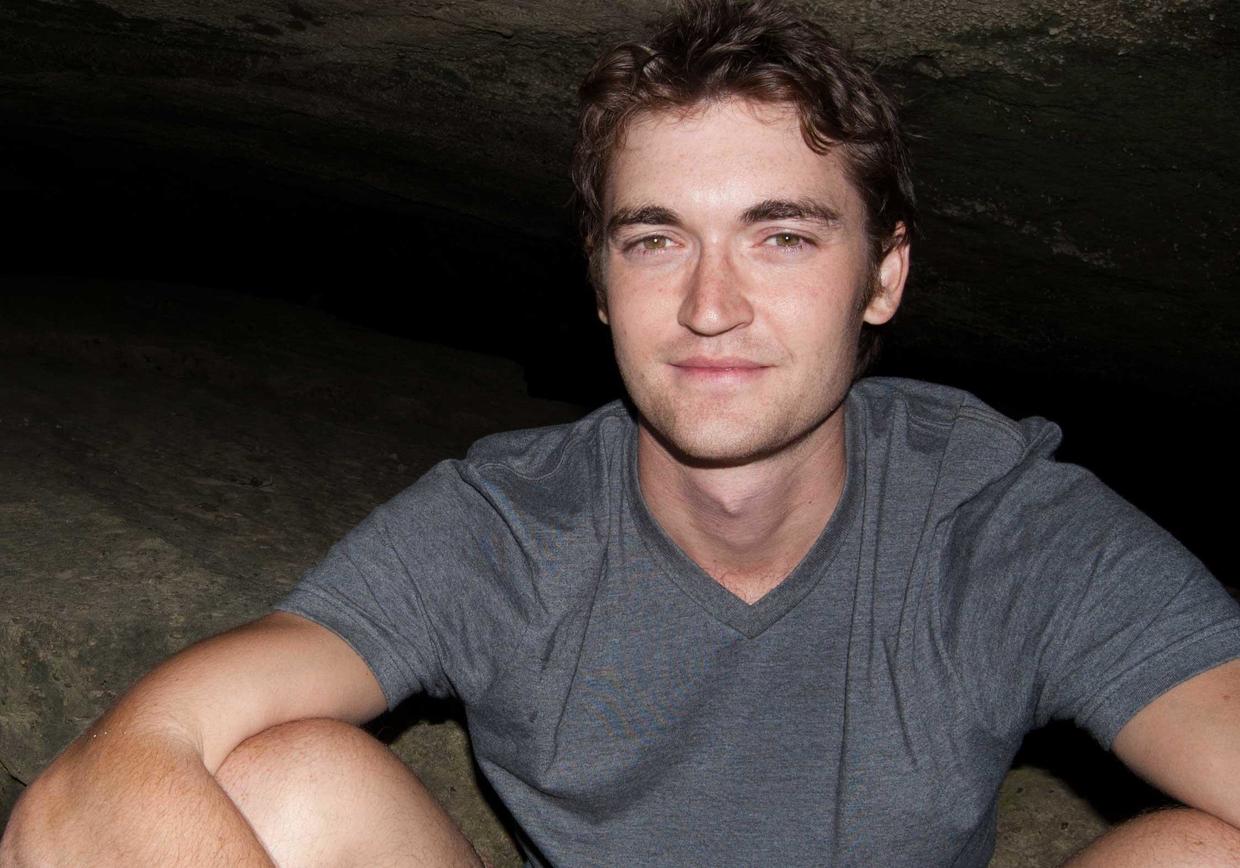 A young man with light brown hair and a gray v-neck shirt sits in a dimly lit cave or overhang
