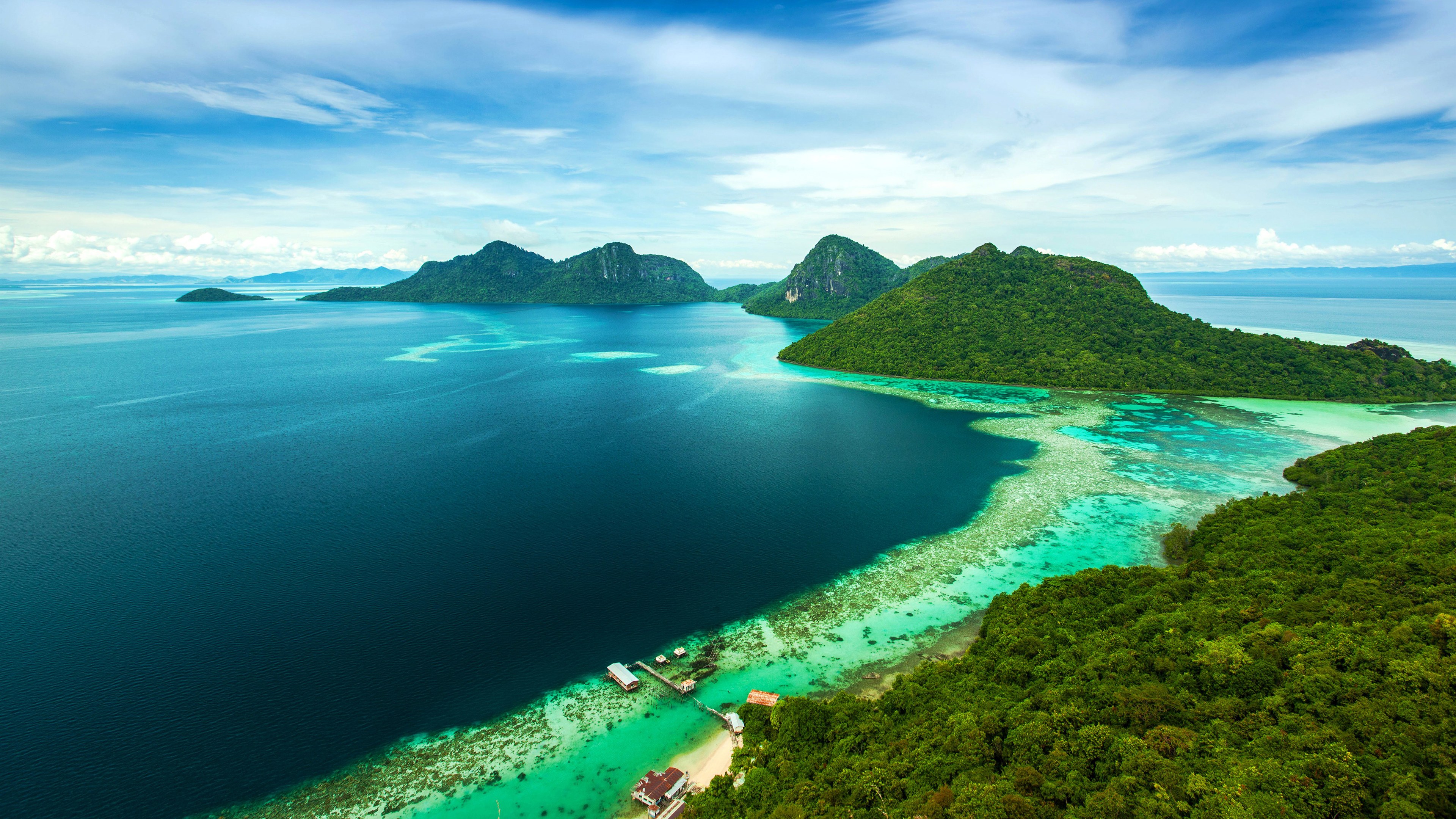 Aerial view of a tropical island with lush green vegetation, surrounded by turquoise water and coral reefs, under a blue sky with white clouds