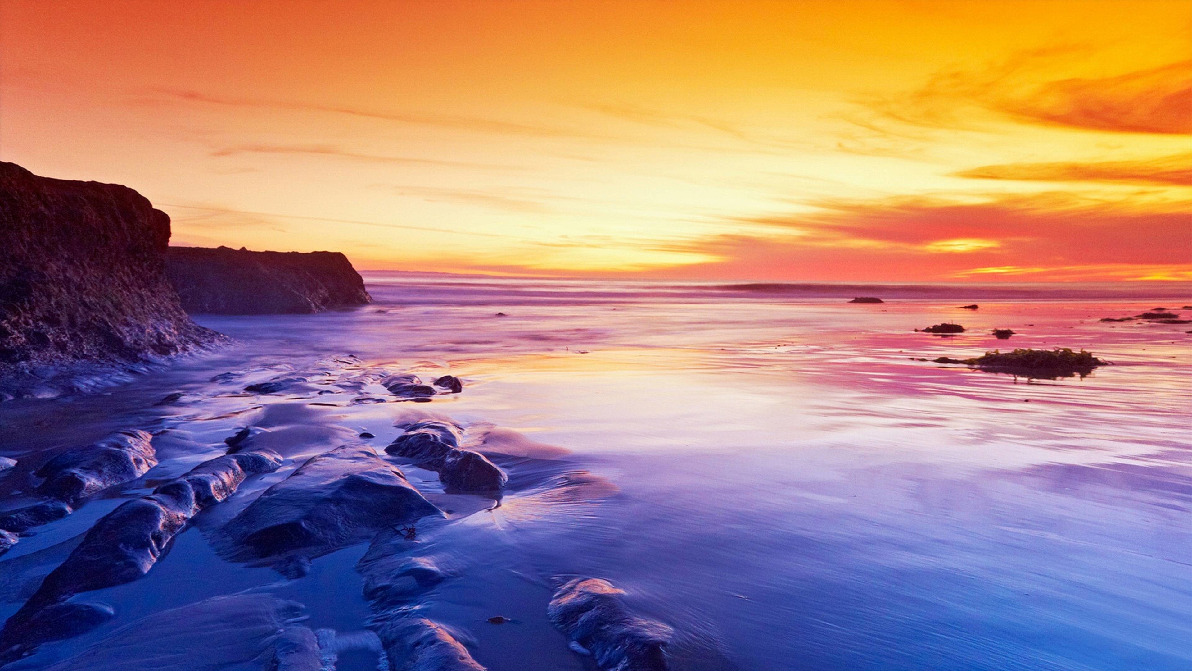 Vibrant sunset over a rocky beach, with the colorful sky reflected in the wet sand and shallow water