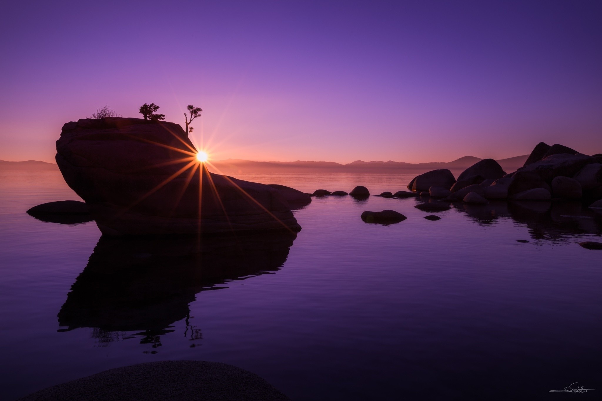 Silhouette of a rock formation in a lake at sunset, with sun rays creating a starburst effect