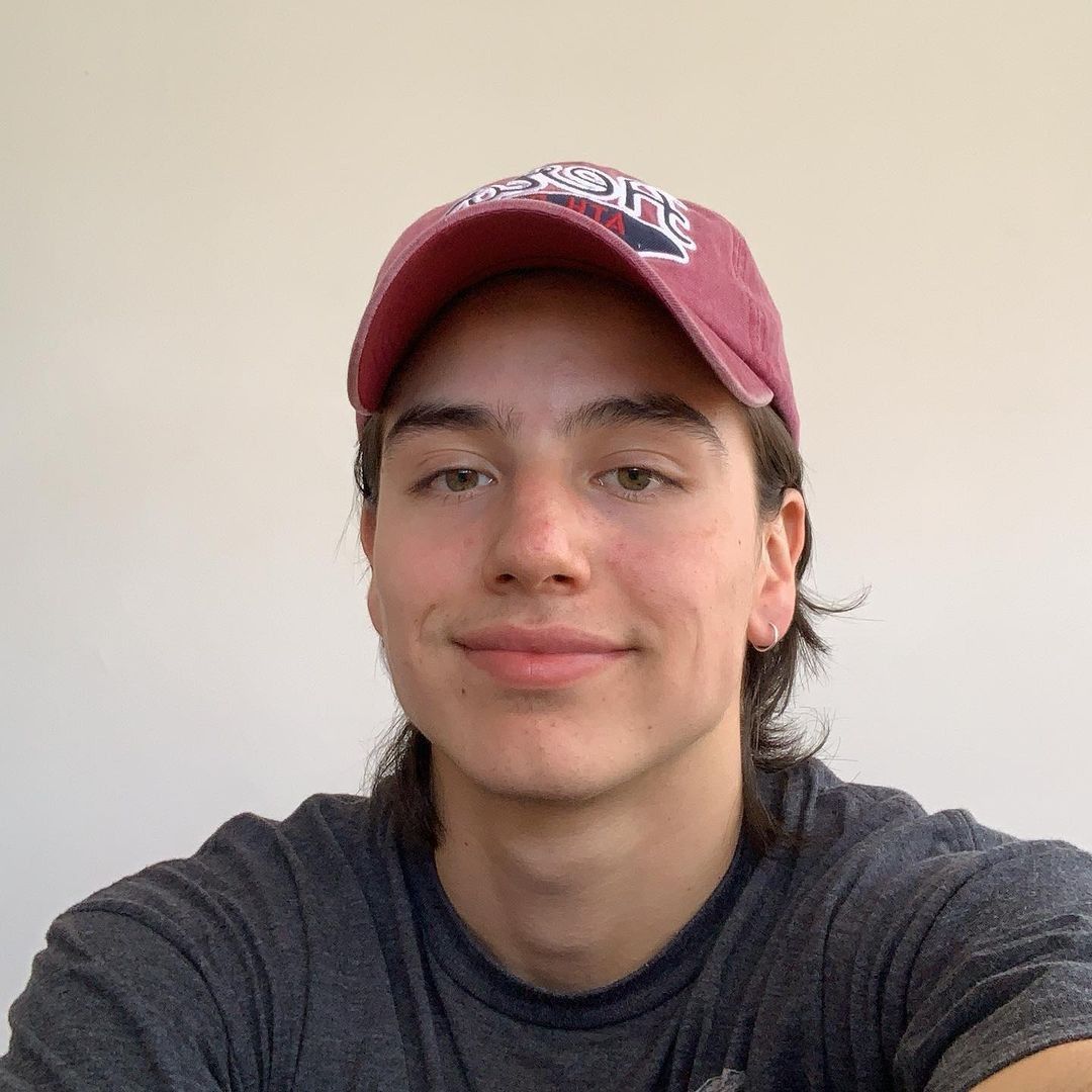 A selfie of a young person with medium-length brown hair wearing a maroon baseball cap and a dark gray t-shirt