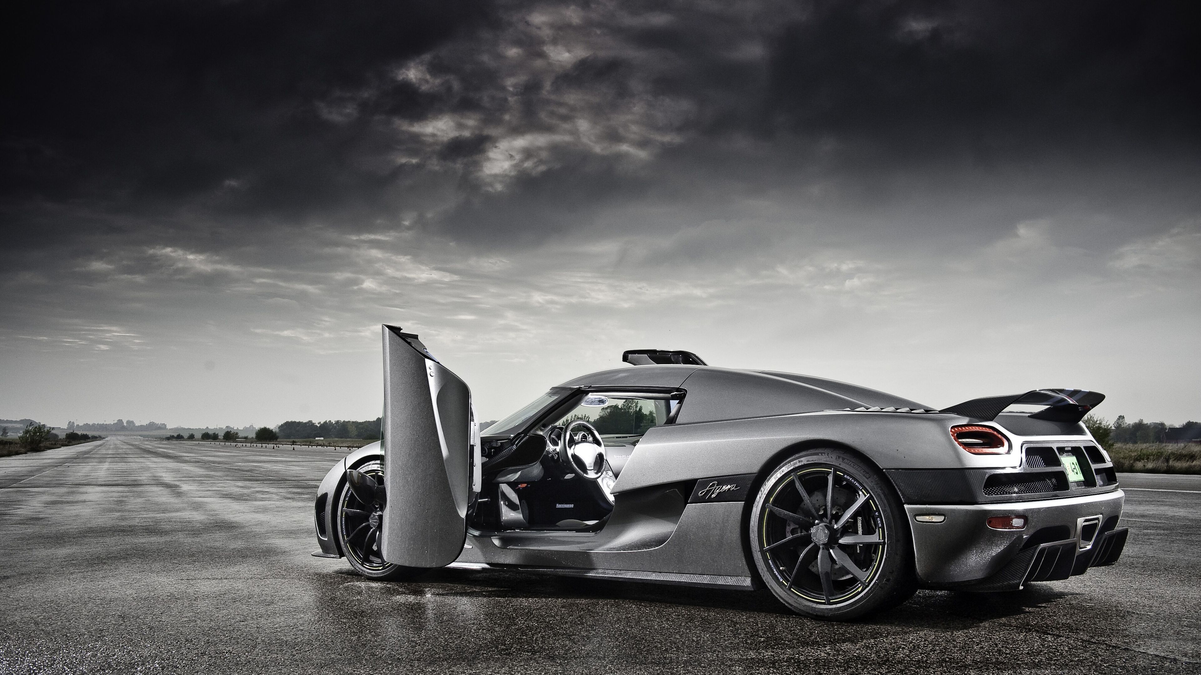 A silver Koenigsegg Agera sports car with its door open, parked on a tarmac under a dramatic cloudy sky