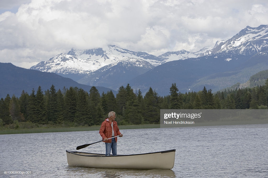 Senior Man Standing In Canoe On River Mountain Range Background
