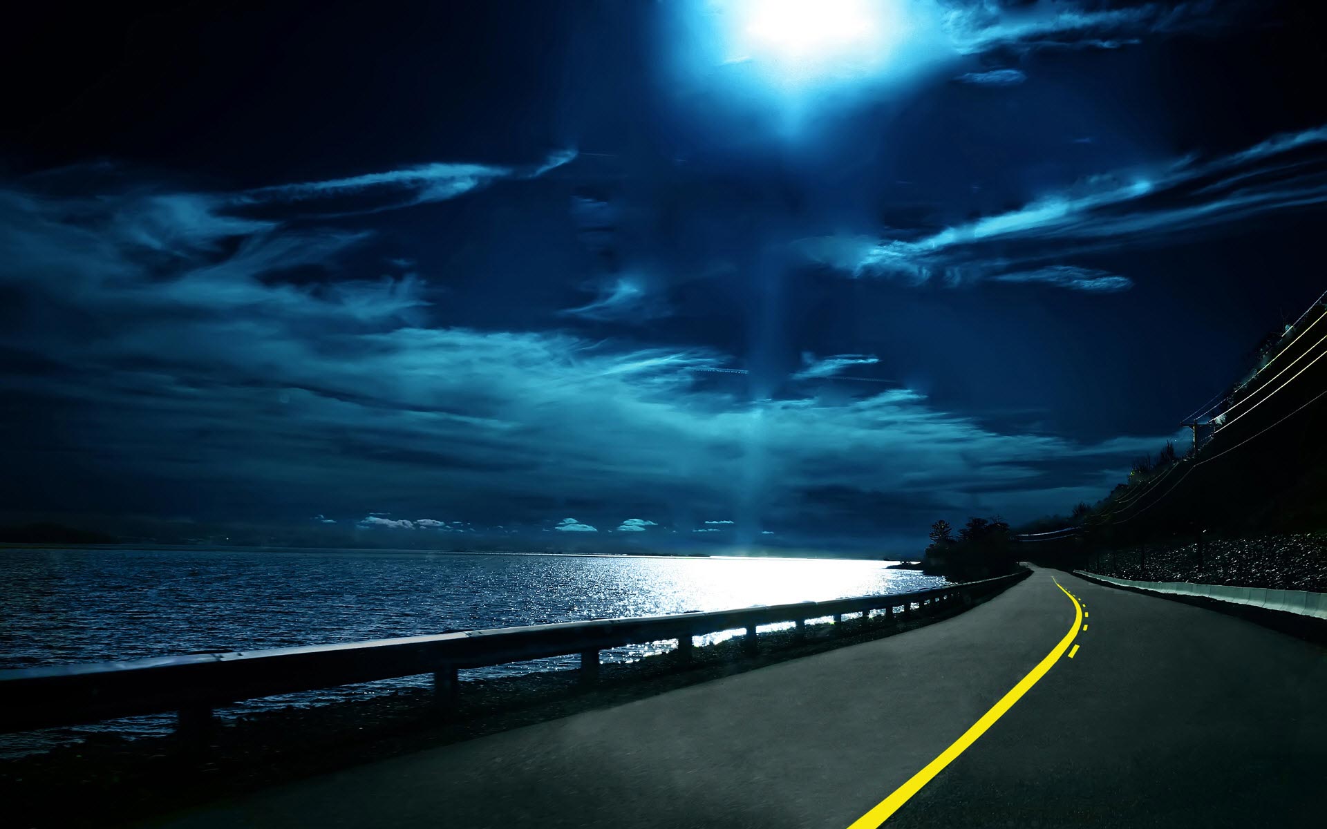 A scenic desktop wallpaper featuring a dark road winding along a coastline under a moonlit sky