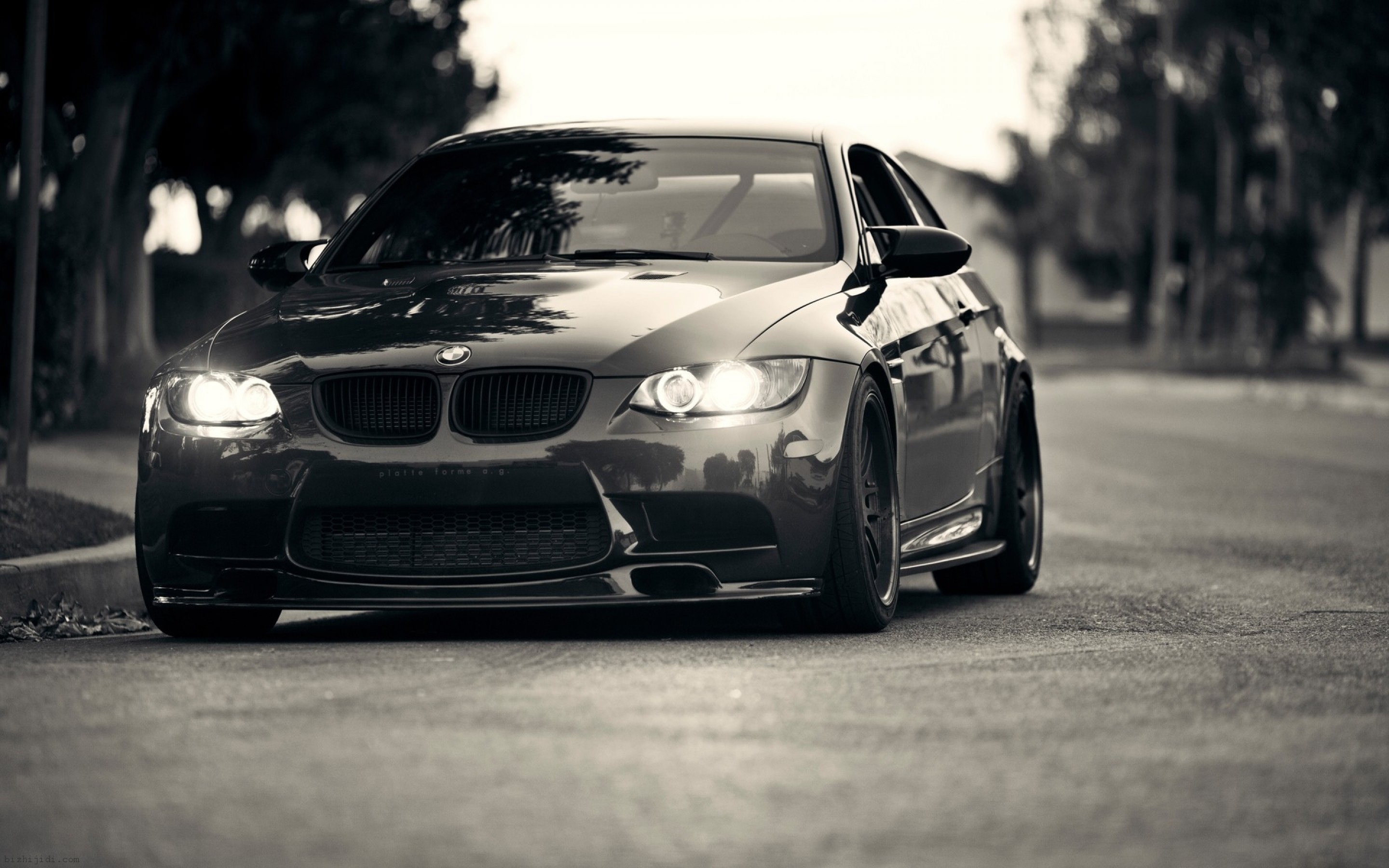 Black and white photo of a BMW M3 parked on a street, headlights on