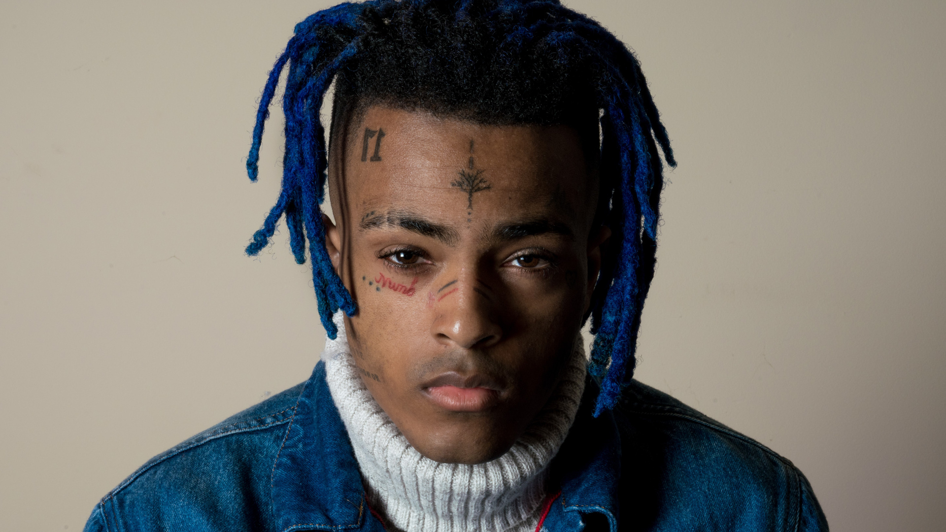 Close-up portrait of XXXTentacion with blue dreadlocks, wearing a denim jacket and a white turtleneck