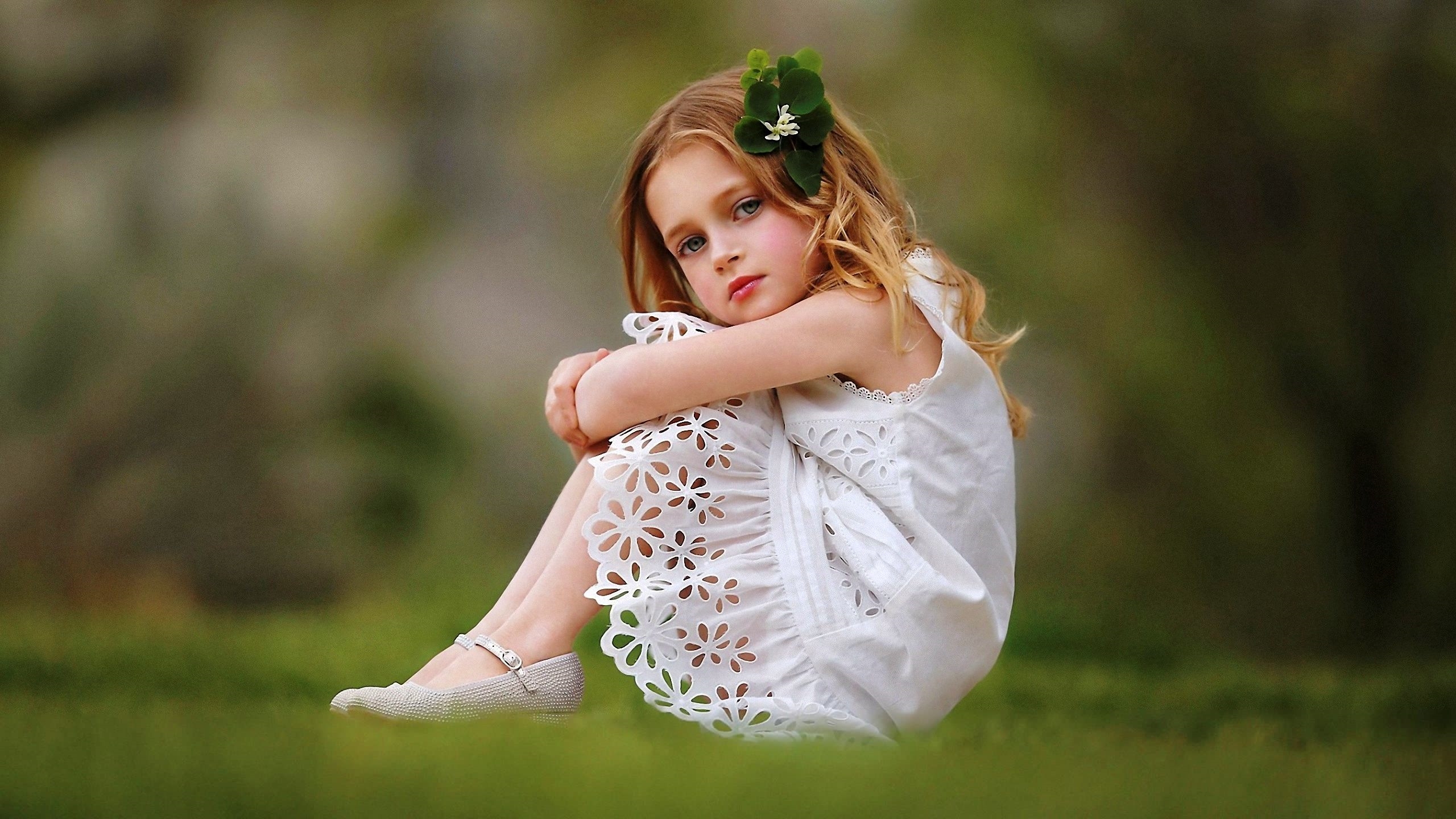 A young girl with blonde hair and a white dress sits on the grass, hugging her knees. A small flower is tucked behind her ear. The background is blurred, suggesting an outdoor setting