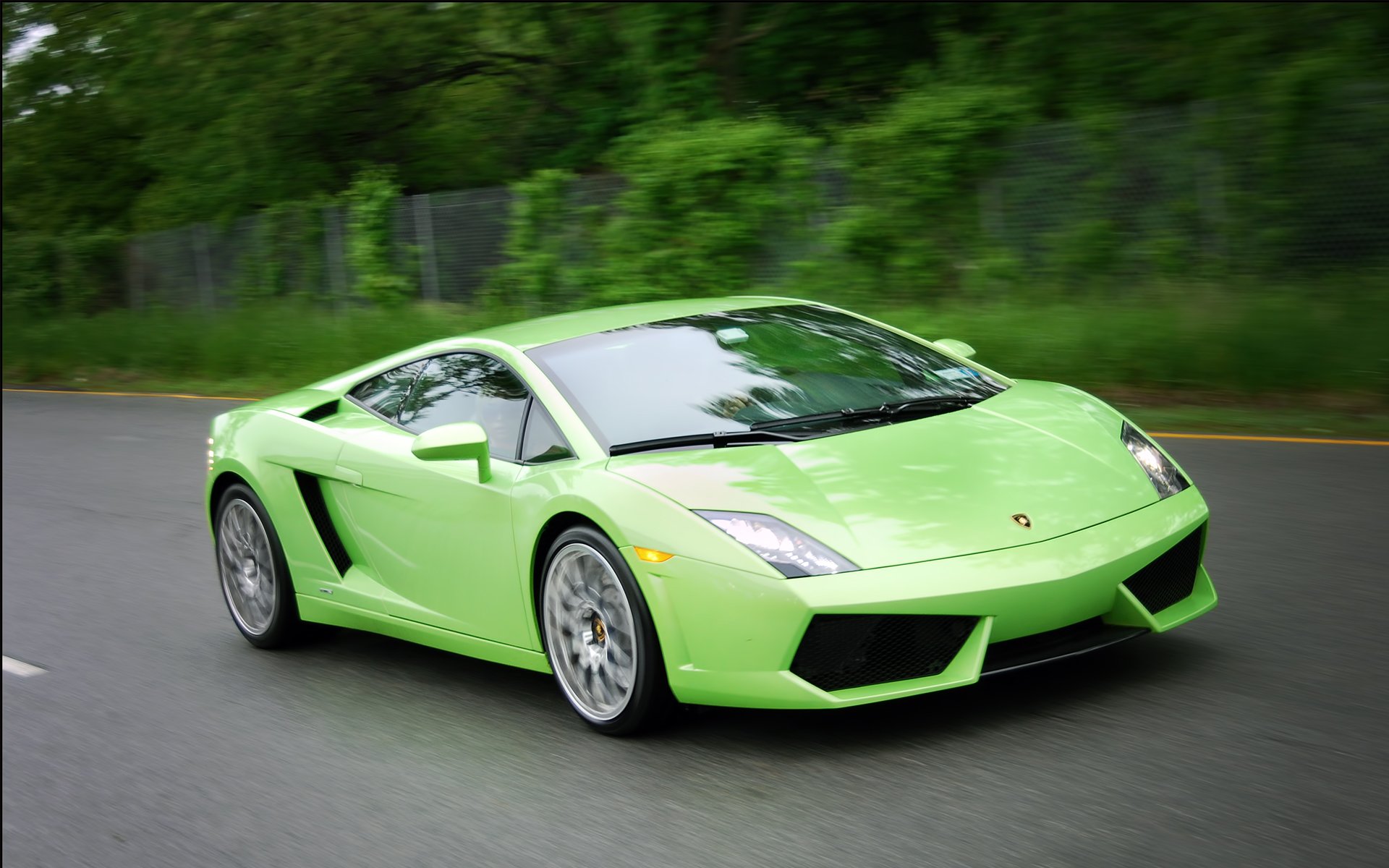 A vibrant green Lamborghini Gallardo speeds down a paved road, blurred background of trees and a fence adds to the sense of motion