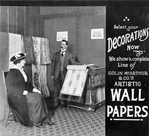 Photograph of a salesman in a wallpaper store showing samples to a