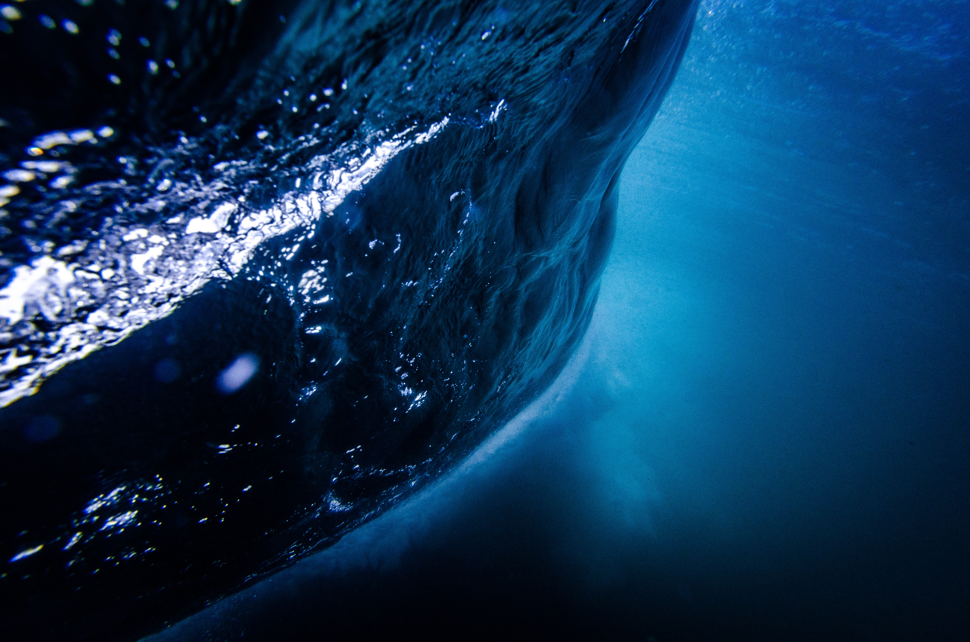 A breathtaking underwater view of a powerful ocean wave, captured in stunning 4K resolution. The deep blue water contrasts with the lighter, bubbly surface of the wave, creating a mesmerizing visual